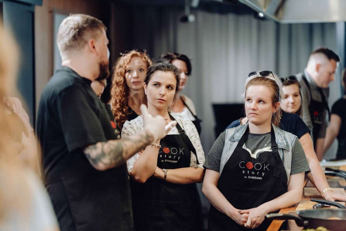 Grupa osób ubranych w czarne fartuchy z logo „Cook Story” stoi w kuchni. Z uwagą przysłuchują się wytatuowanemu mężczyźnie, który coś wyjaśnia. Na otaczających je stołach widać przybory kuchenne i składniki, co doskonale uchwycono na tej fotografii eventowej.