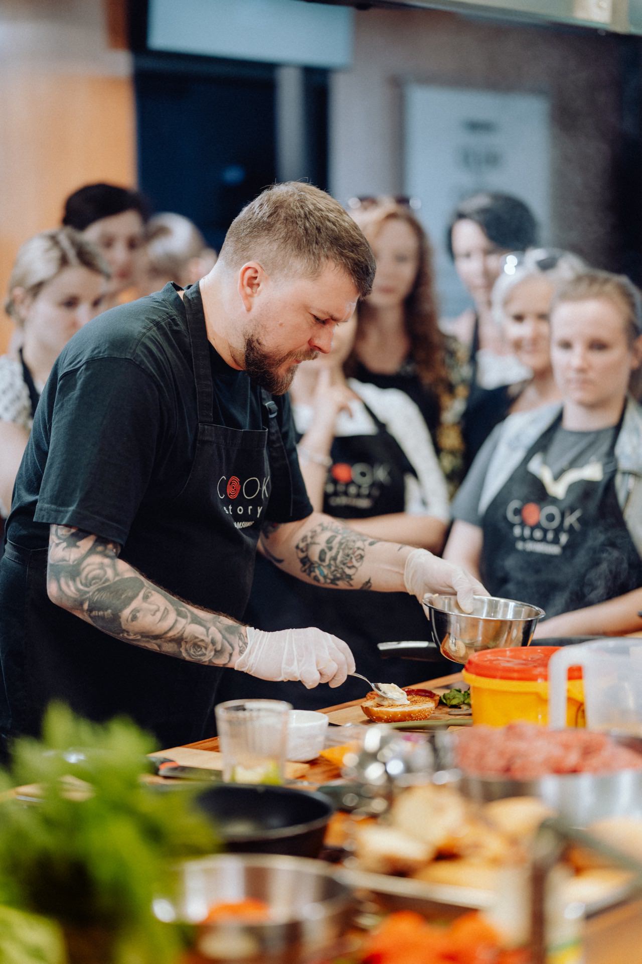 A chef with tattoos and a beard, wearing a black T-shirt with the word "COOK" on it, prepares food while an attentive audience watches. In this photo essay of the event, he works in a kitchen full of various ingredients and utensils, wearing white gloves for hygiene. 