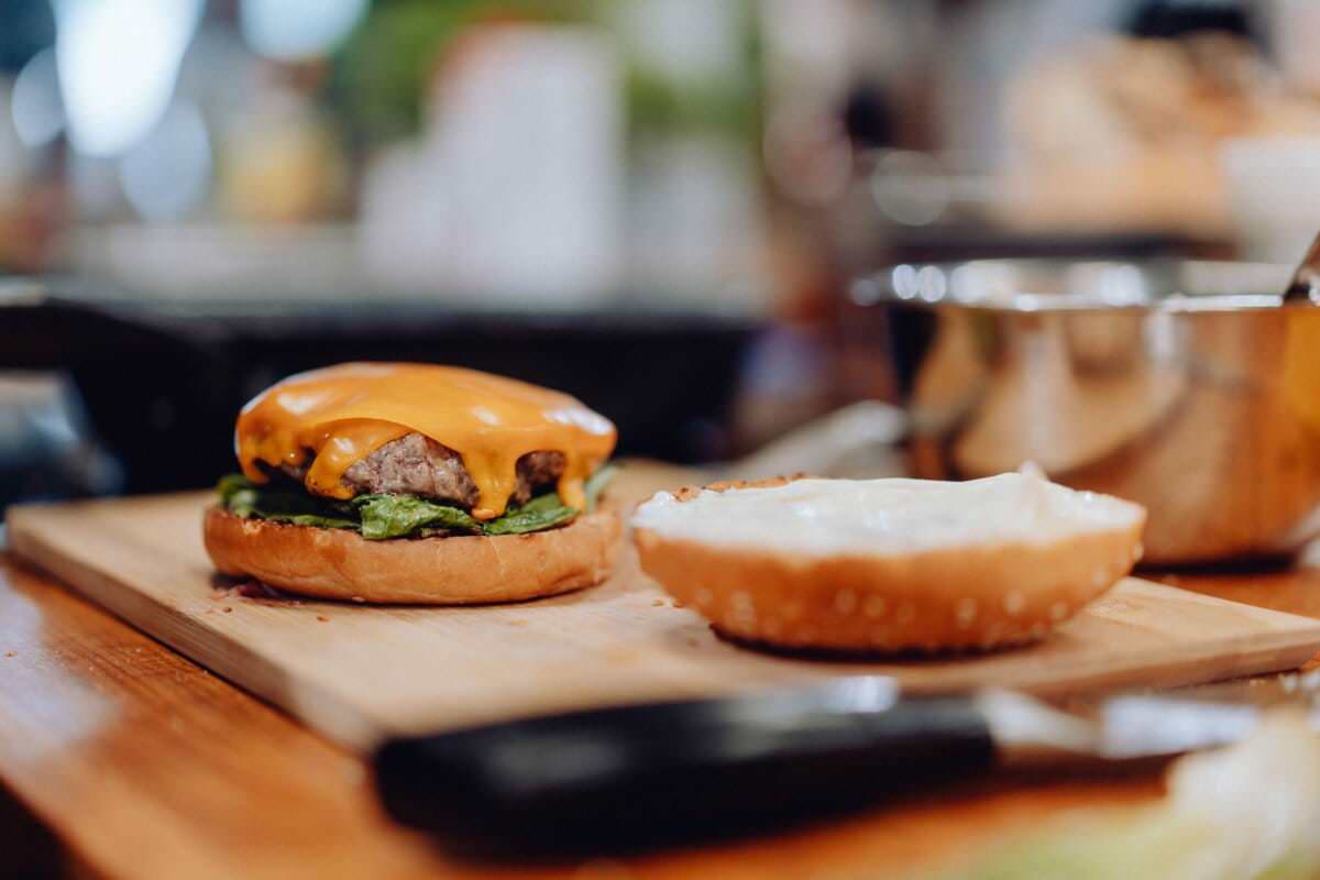 Zbliżenie częściowo złożonego cheeseburgera na drewnianej desce do krojenia. Pasztecik burgerowy posypany jest topionym serem i sałatą. Obok kładzie się górną połowę bułki z majonezem. W pobliżu widać nóż kuchenny i mały garnek, przypominające detale znalezione na fotografiach fotorelacja z wydarzeń.