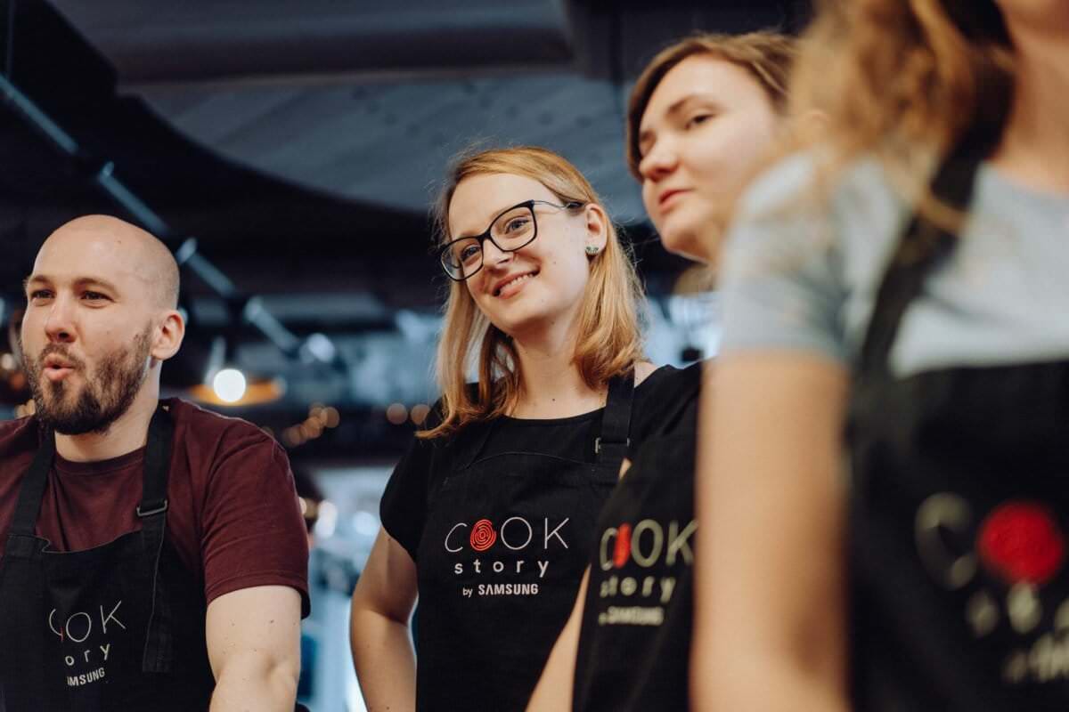Cztery osoby stoją razem, nosząc czarne fartuchy z napisem „Cook Story by Samsung”. Jedna osoba w środku uśmiecha się i nosi okulary, podczas gdy inni mają neutralne lub skupione wyrazy twarzy. Wyglądają, jakby byli w kuchni, uchwyceni na fotografii eventowej, która podkreśla ten moment.
