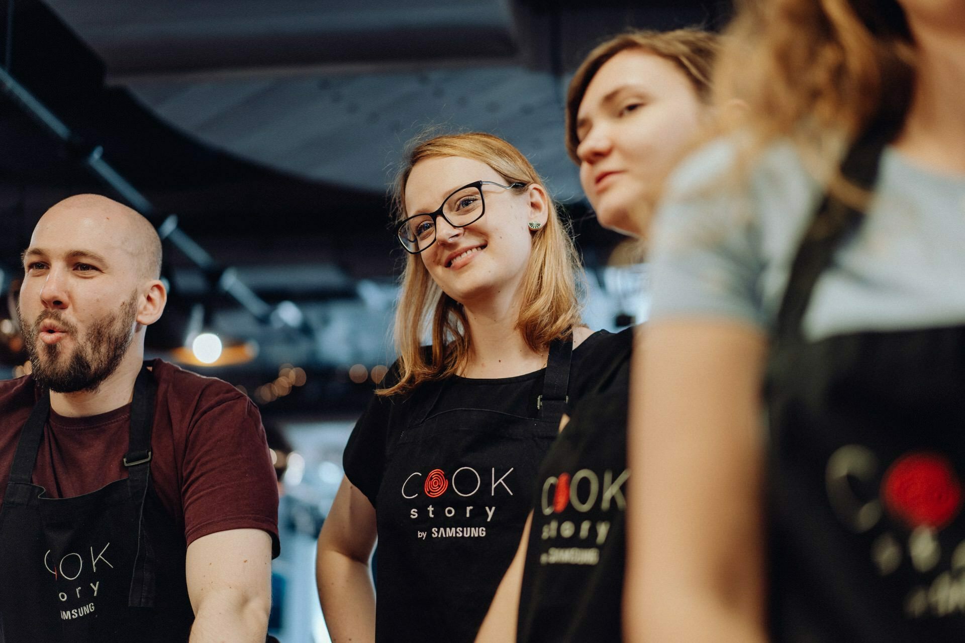 Four people stand together, wearing black aprons that read "Cook Story by Samsung." One person in the middle is smiling and wearing glasses, while the others have neutral or focused expressions on their faces. They look as if they are in the kitchen, captured in an event photograph that highlights the moment.  