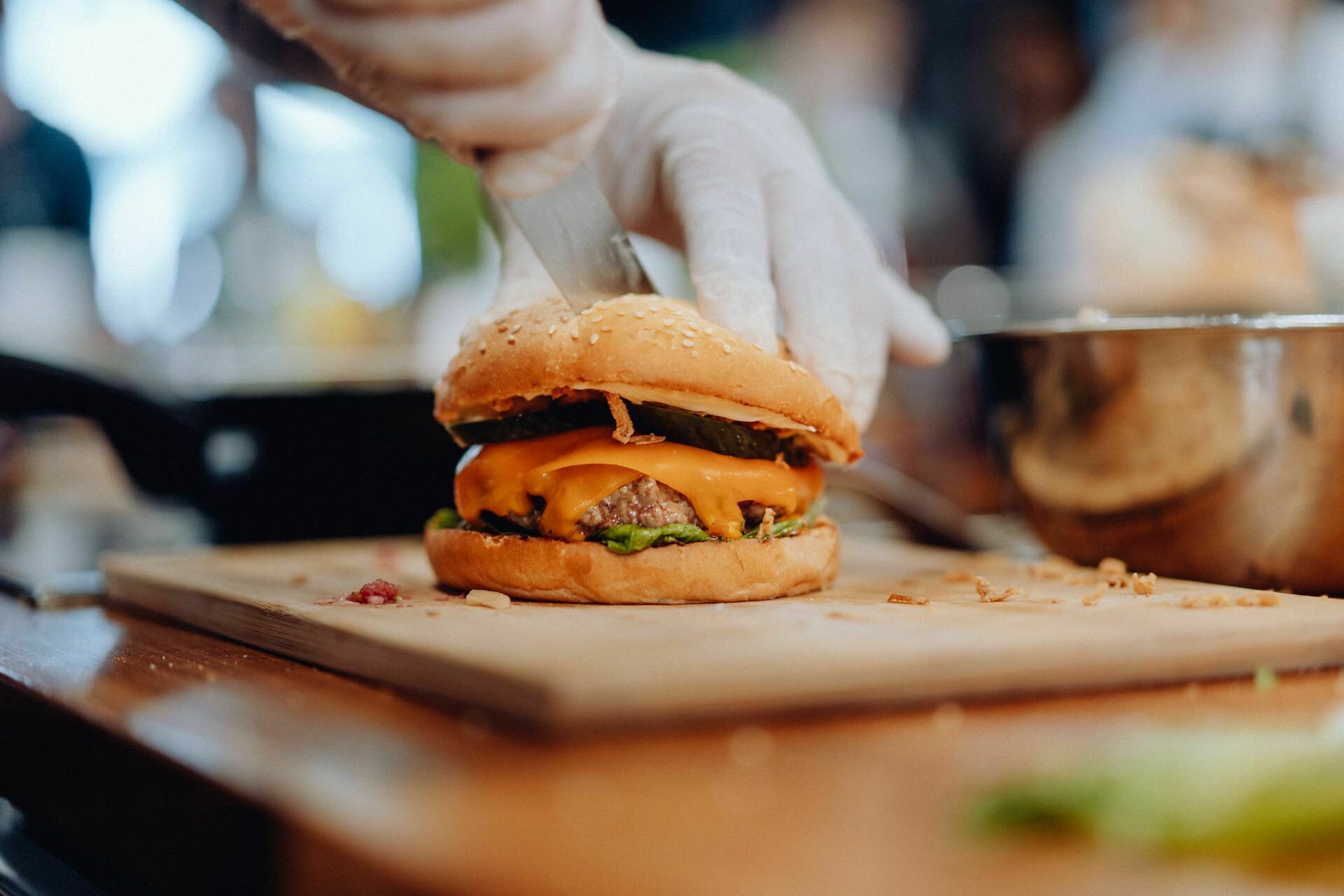 A person wearing gloves prepares a cheeseburger with lettuce, beef pate, melted cheddar cheese and pickle slices in a sesame seed bun. The burger stands on a wooden countertop in a crockery - perfect for event photography showcasing culinary skills. 