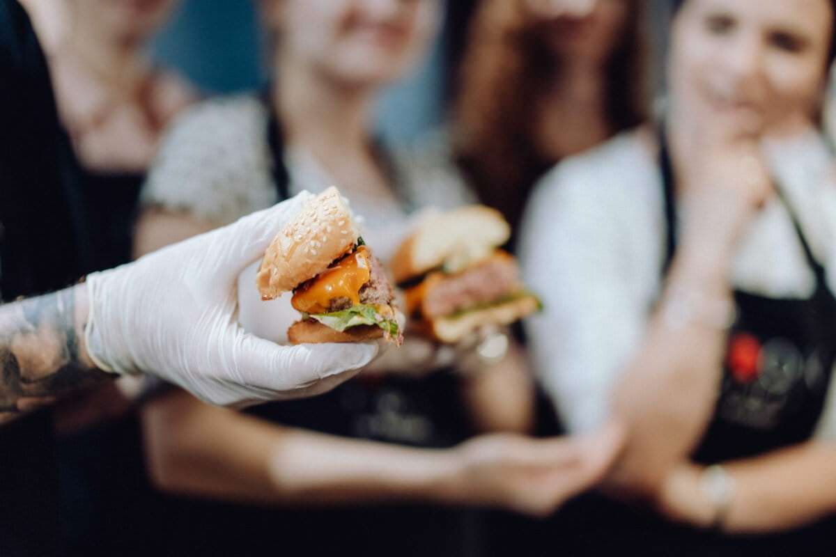 Osoba w rękawiczce trzyma dwa burgery z bułką sezamową, każdy wypełniony pasztecikami wołowymi, serem i sałatą. W tle z uwagą przygląda się grupa osób w czarnych fartuchach – doskonały moment na fotografię eventową.