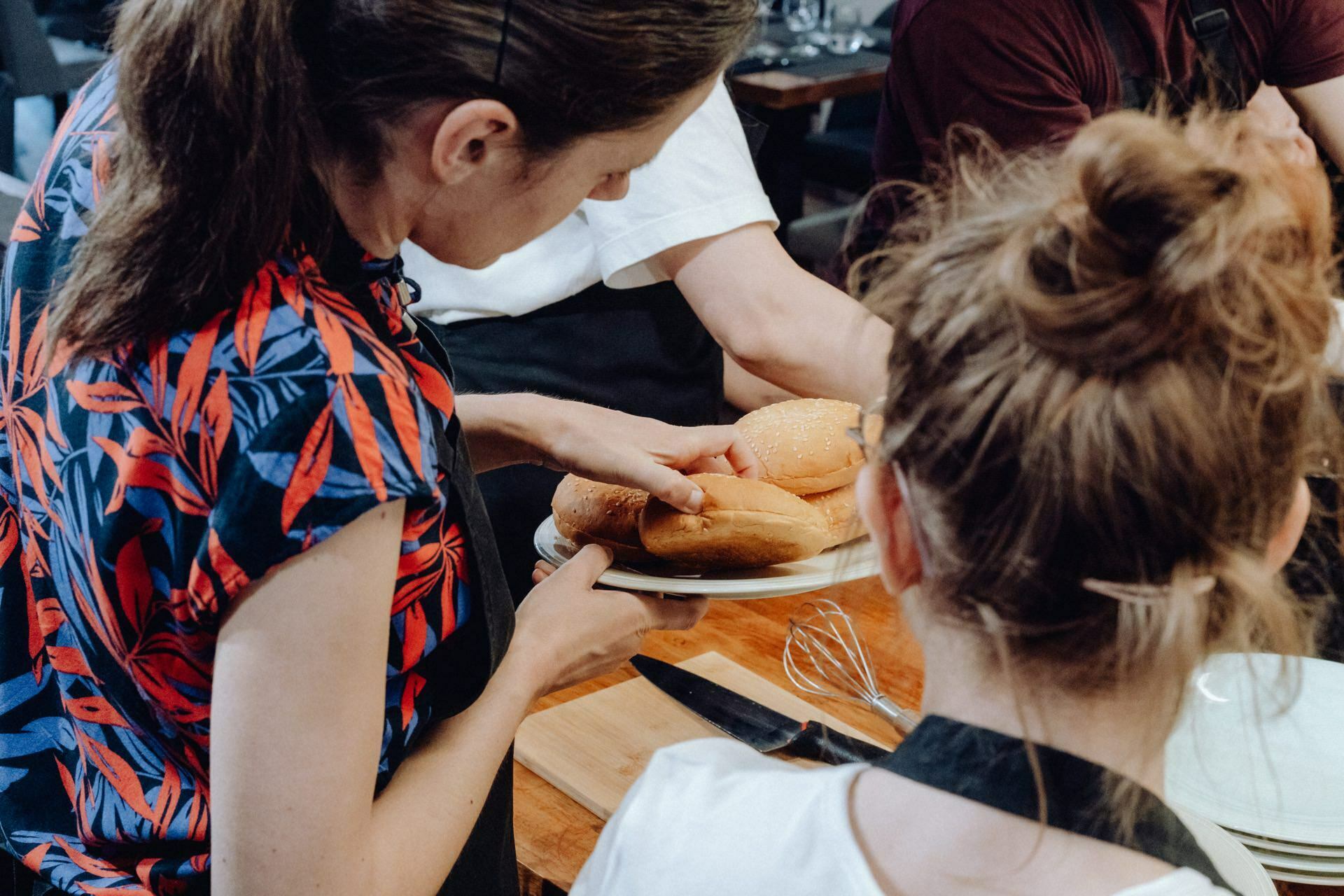 Two people can be seen from behind at the table: one is putting rolls on a plate and the other is watching. Both have their hair up and are wearing aprons. Also visible on the table is a set of kitchen utensils and a cutting board - a perfect moment captured in an event photograph.  