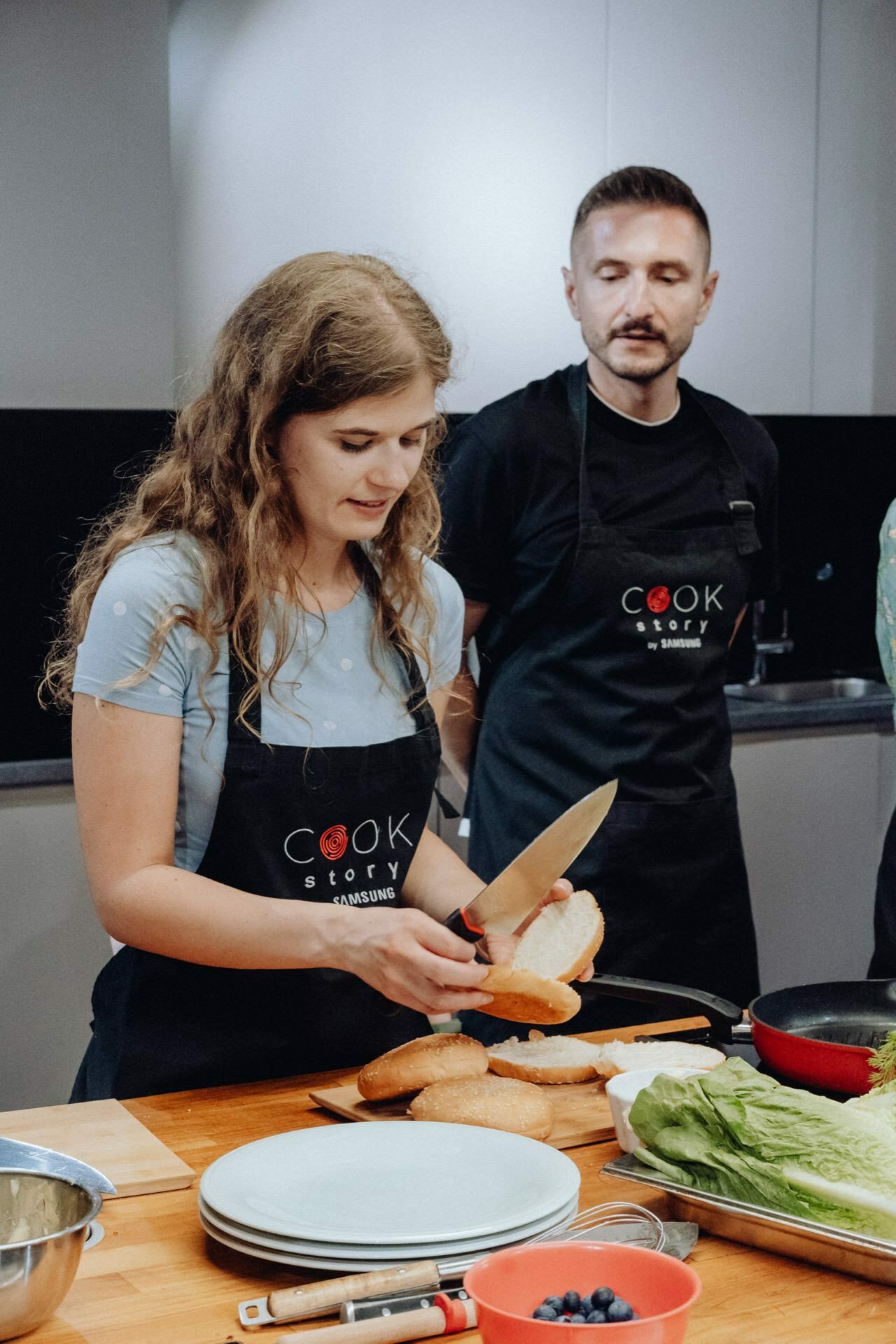 A woman slices a baguette in a black apron with the words "Cook Story by Samsung" written on it. She stands at the kitchen counter with plates, lettuce and other ingredients, creating the perfect scene for an event photographer. A man in a similar apron watches her in the background.  