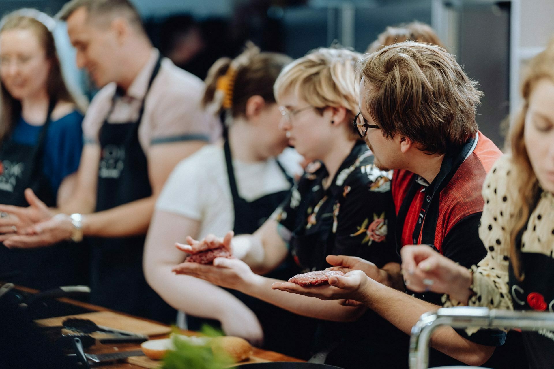 A group of people dressed in aprons are gathered around a wooden table, busy cooking. Two people in the foreground are shaping raw ground meat with their hands, while others are watching or participating. The background is blurred with kitchen equipment, creating the perfect setting for a photo-reportage of the events.  