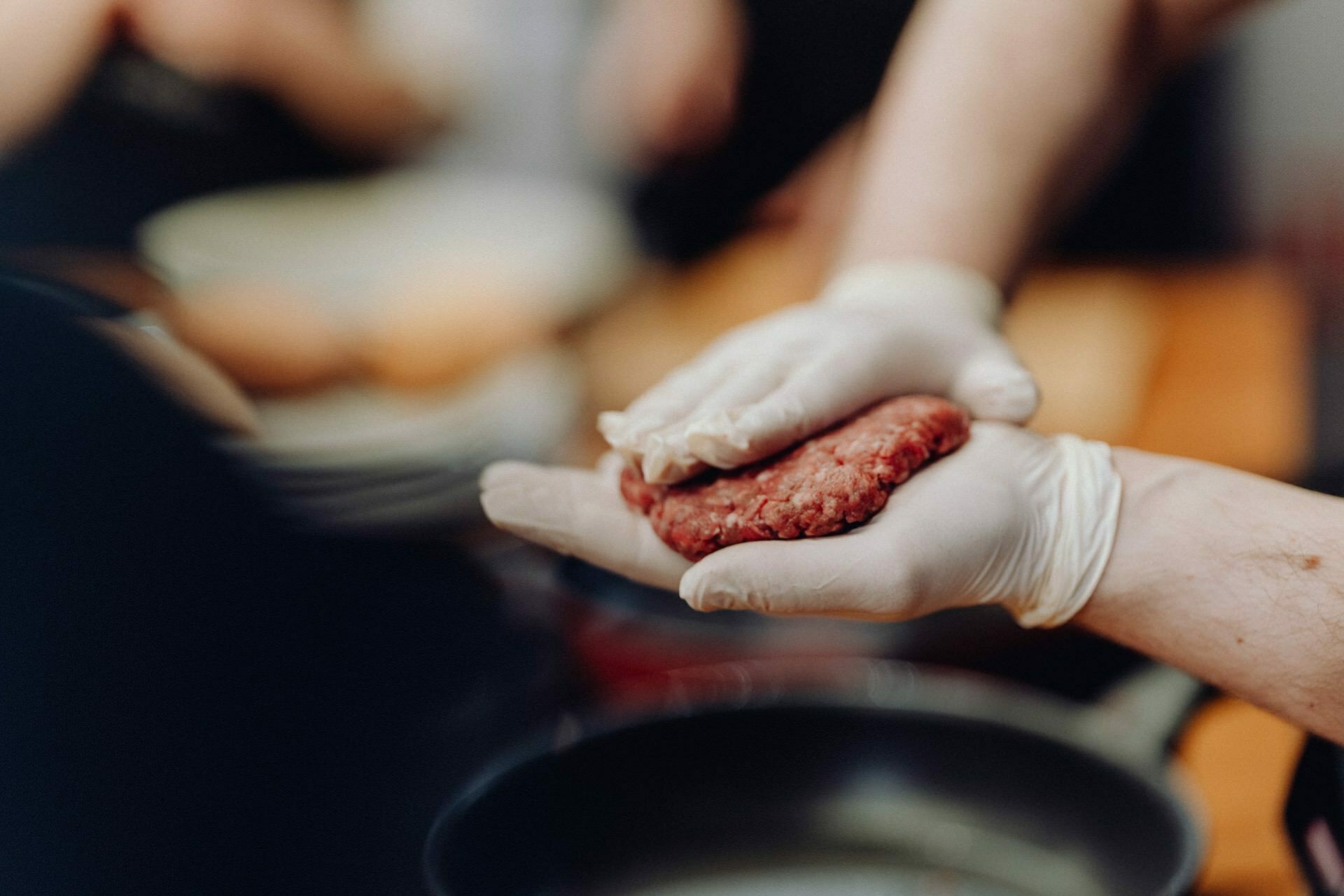 A person wearing white gloves forms a raw hamburger in his hands over a kitchen countertop. Kitchen utensils and ingredients are blurred in the background, capturing a candid moment reminiscent of event photography. 