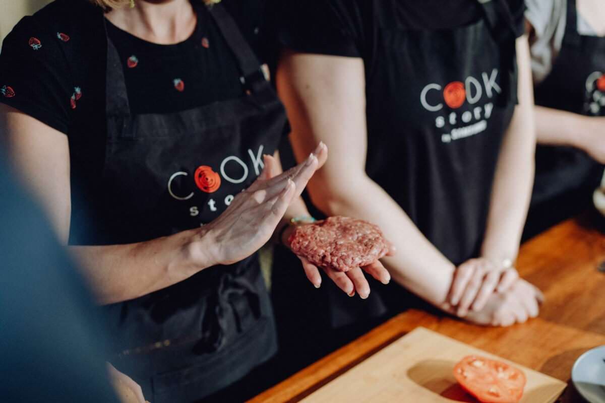 Trzy osoby biorą udział w lekcjach gotowania. Osoba na pierwszym planie formuje rękami surowego hamburgera. Wszyscy mają na sobie czarne fartuchy z nadrukiem „Cook Story”. W tle na drewnianym blacie leży pokrojony pomidor, idealny na fotografię eventową.