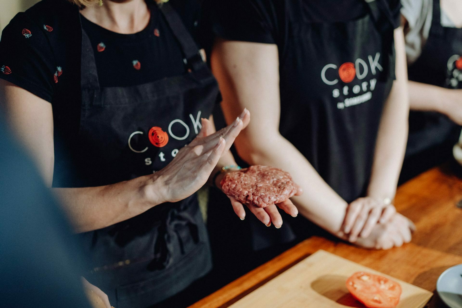 Three people take part in a cooking class. The person in the foreground is forming a raw hamburger with his hands. They are all wearing black aprons with "Cook Story" printed on them. In the background, a sliced tomato lies on a wooden countertop, perfect for event photography.   