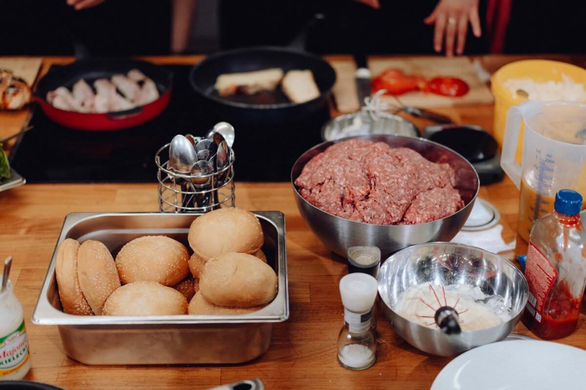 Blat kuchenny wypełniony składnikami i przyborami do robienia burgerów przypomina scenę z fotografii eventowej. Na tacy znajdują się bułki burgerowe, miska surowego mięsa mielonego, pokrojone w plasterki pomidory, plastry sera, różne przyprawy, shakery do przypraw i patelnia, na której gotują się kotleciki hamburgerowe.