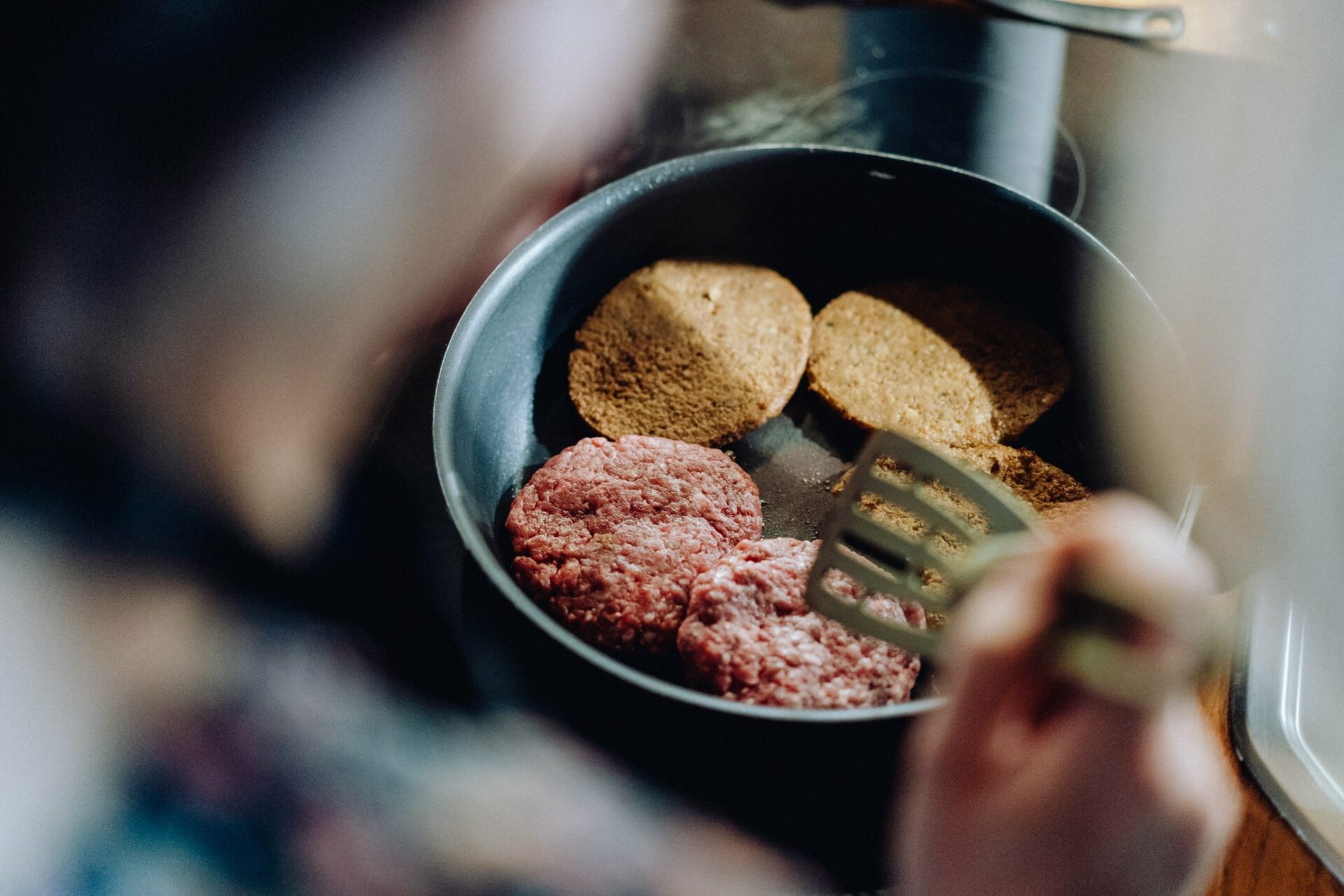 A person cooking pasties with ground meat and bread rolls in a pan on a cooktop, holding a spatula over the food. The photo, taken from an over-the-shoulder perspective, captures the essence of event photography's focus on the cooking process. 