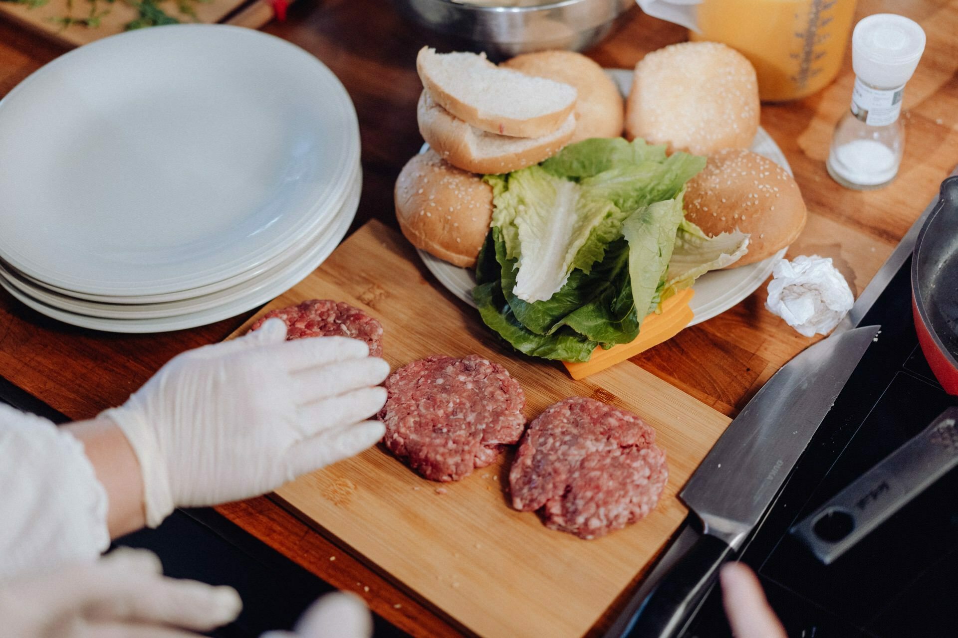 A person wearing gloves prepares raw hamburgers on a wooden cutting board. Nearby are buns, lettuce, cheese, a stack of plates, a knife and other kitchen utensils. A measuring cup and salt shaker are visible in the background - a perfect shot for an event shoot.  