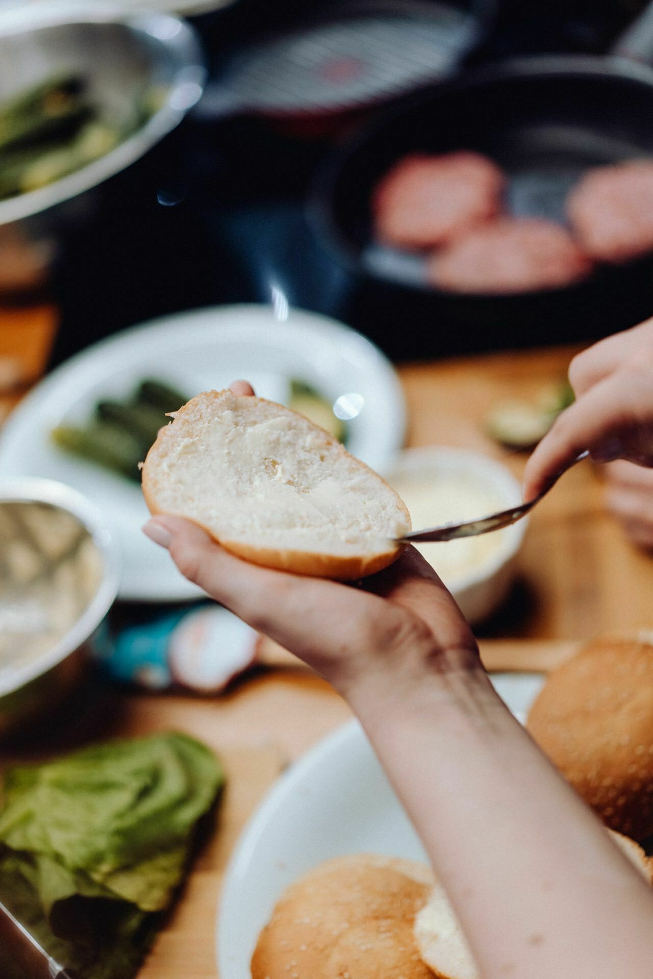 In the kitchen, a person spreads condiment on a burger bun cut in half with a knife. Various ingredients, including lettuce, pickles and raw burger patties, are arranged on plates and bowls on a countertop in the background. This scene could be part of a photo essay of the event captured by an event photographer warsaw.  