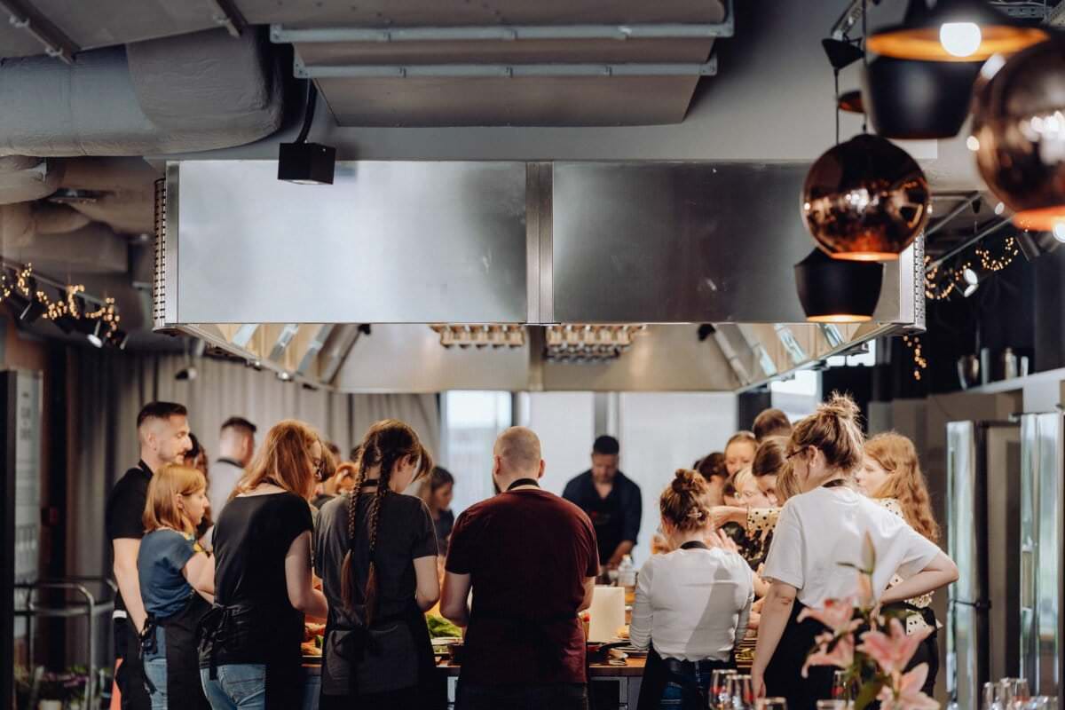 Grupa osób gromadzi się w nowoczesnej kuchni w stylu industrialnym. Zajmują się różnymi czynnościami kulinarnymi wokół dużej centralnej wyspy. Przestrzeń jest dobrze oświetlona, wyposażona w okapy wentylacyjne, oprawy oświetleniowe z błyszczącej miedzi i duże okna w tle – idealne na fotorelację z wydarzeń.