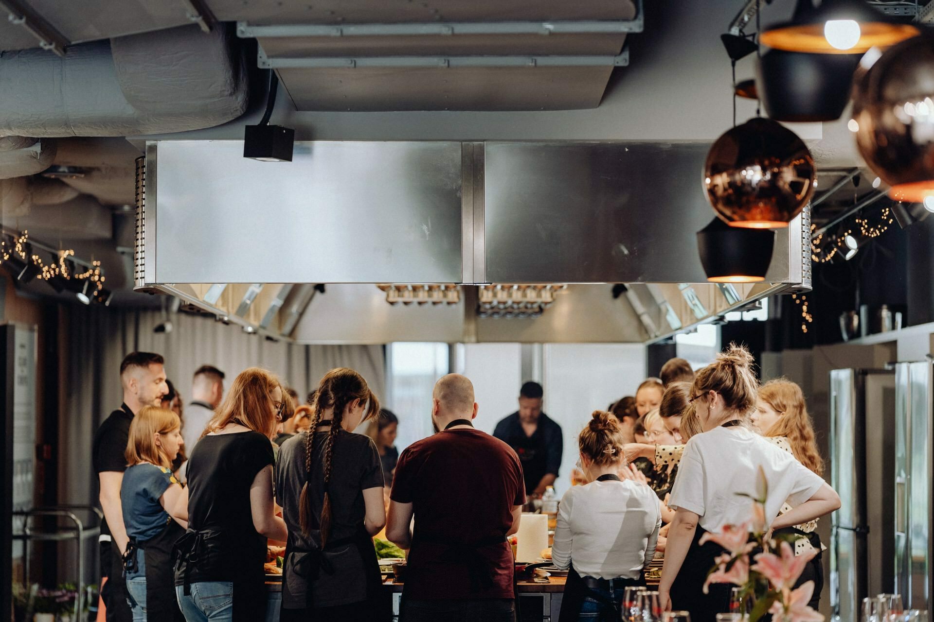 A group of people gather in a modern industrial-style kitchen. They engage in various cooking activities around a large central island. The space is well-lit, with ventilation hoods, shiny copper light fixtures and large windows in the background - perfect for photo opportunities of the events.  