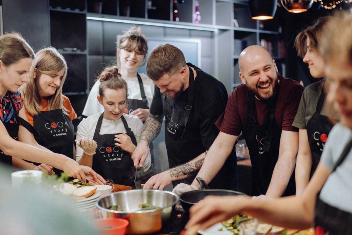 Grupa ośmiu uśmiechniętych osób ubranych w fartuchy gromadzi się wokół wyspy kuchennej i wspólnie przygotowuje jedzenie. Sceneria przypomina lekcję gotowania lub wspólne gotowanie z różnymi miskami i składnikami na blacie, dzięki czemu idealnie nadaje się do fotorelacji z wydarzeń.