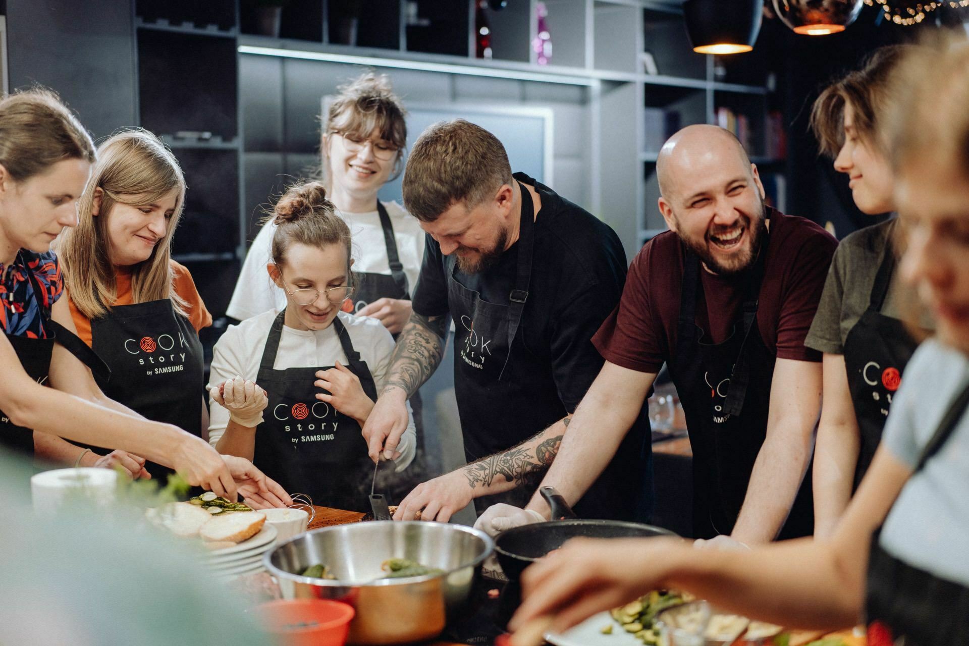 A group of eight smiling people dressed in aprons gather around a kitchen island and prepare food together. The scenery is reminiscent of a cooking lesson or communal cooking with various bowls and ingredients on the countertop, making it ideal for photo-reporting events. 