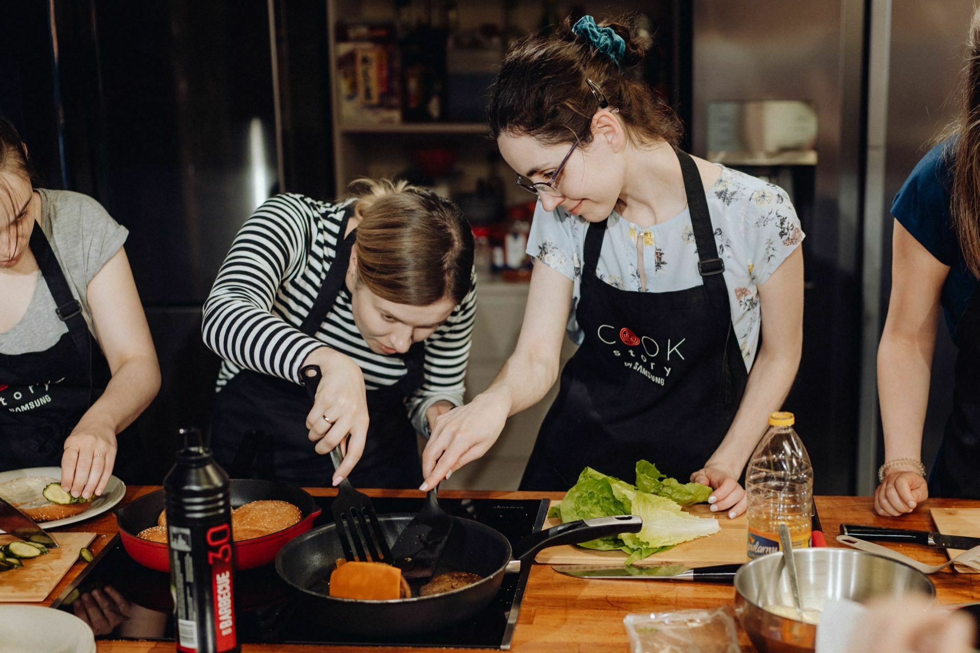 Four people in aprons gather around the kitchen island and cook. Two women in the middle focus on flipping a burger in a skillet. Various ingredients and utensils are spread out on the countertop. In the background you can see a modern kitchen setup, perfect for event photography in Warsaw.   