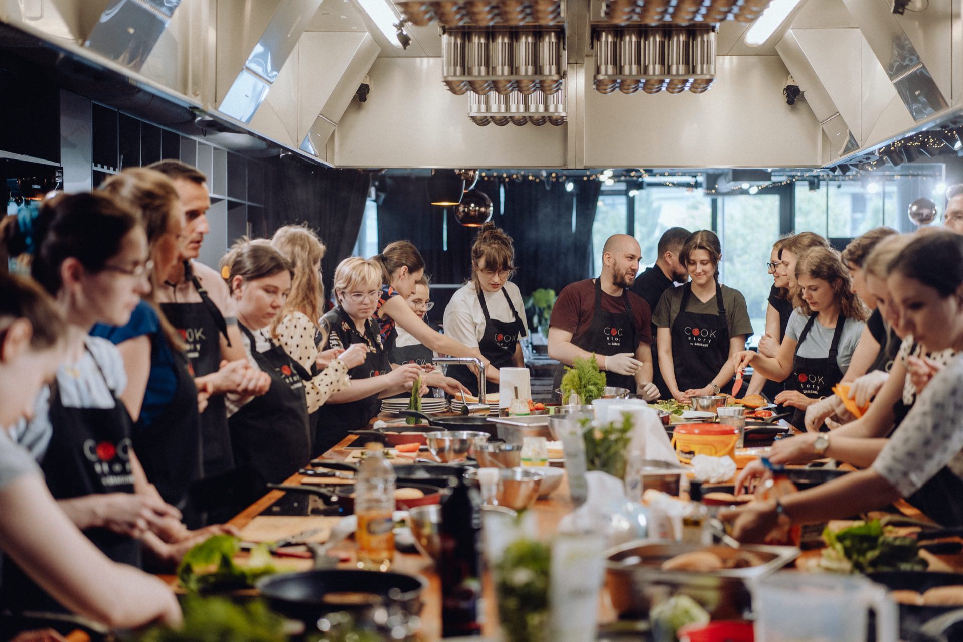 A large group of people dressed in aprons and taking cooking classes gather in a modern kitchen. They work together at a long countertop filled with various ingredients, utensils and kitchenware. The atmosphere seems lively and cooperative, perfect for an immersive photo shoot.  