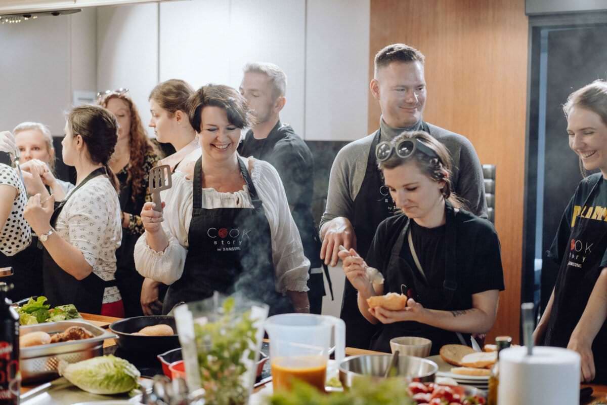 Grupa ludzi zebrała się w kuchni i wzięła udział w lekcjach gotowania. Mają na sobie czarne fartuchy, niektóre z napisem „Akademia Kucharska”. Na ladach porozrzucane są różne składniki i przybory kuchenne, a wszyscy wydają się być uśmiechnięci i zadowoleni z zajęć – idealne miejsce na fotografię eventową.