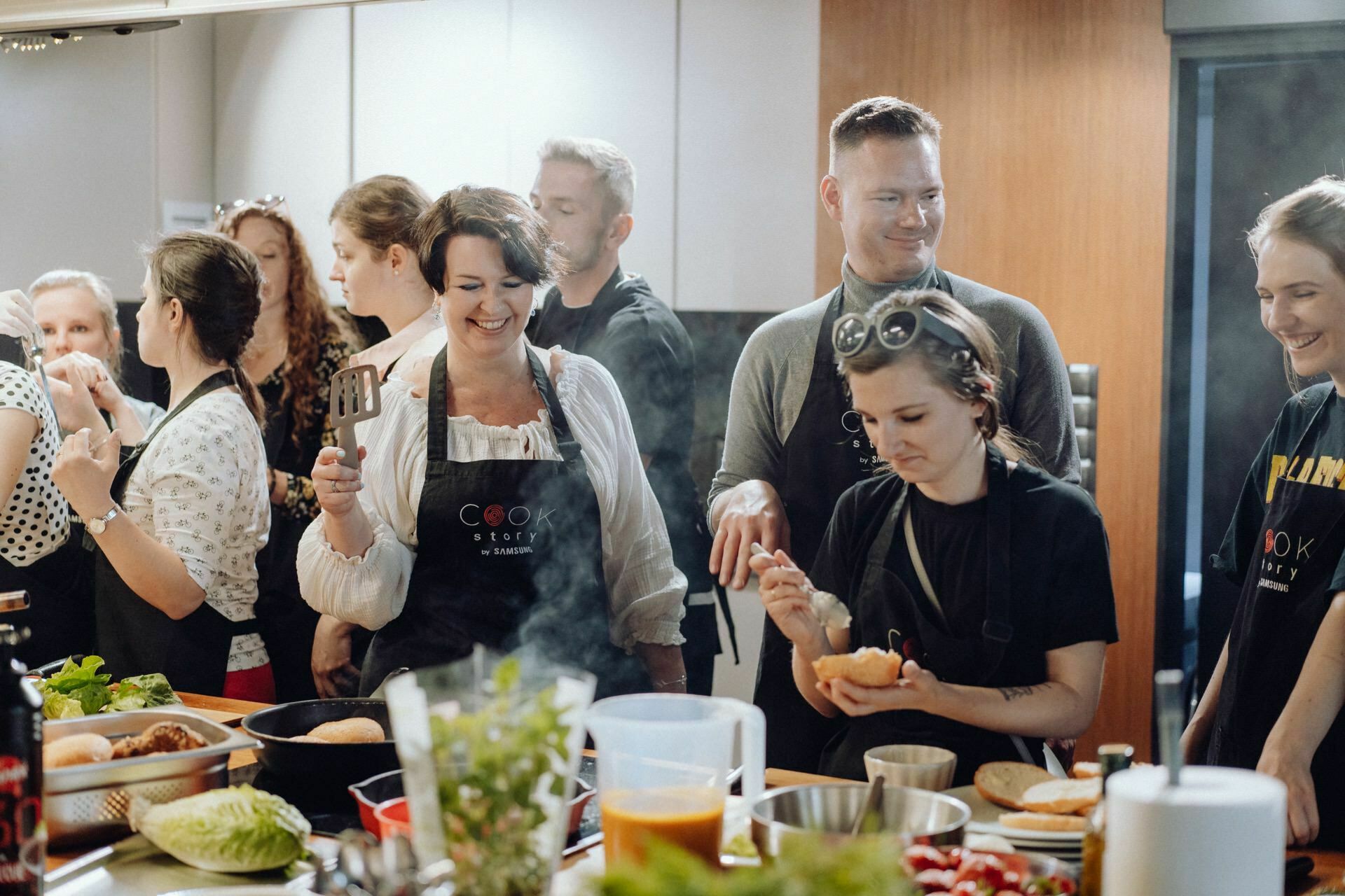 A group of people have gathered in the kitchen and taken cooking lessons. They are wearing black aprons, some with the words "Cooking Academy" written on them. Various ingredients and cooking utensils are scattered on the counters, and everyone seems to be smiling and enjoying the class - a perfect place for event photography.  