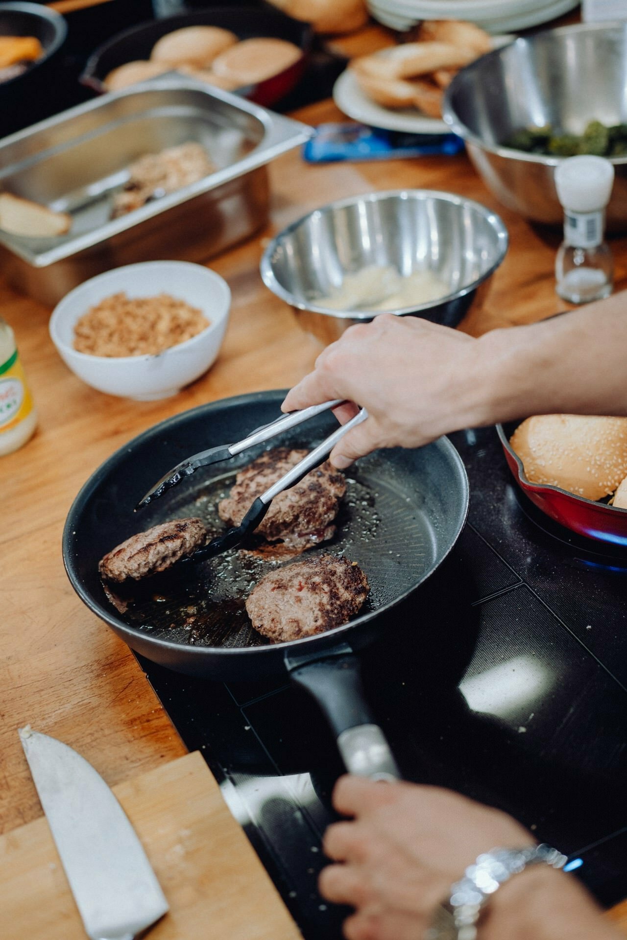 Close-up of a person cooking hamburgers in a pan on a cooktop. The person is using tongs to flip the patties. Various ingredients and kitchen utensils are visible in the background, including bowls, a knife and buns - a perfect scene for event photography capturing culinary moments.  