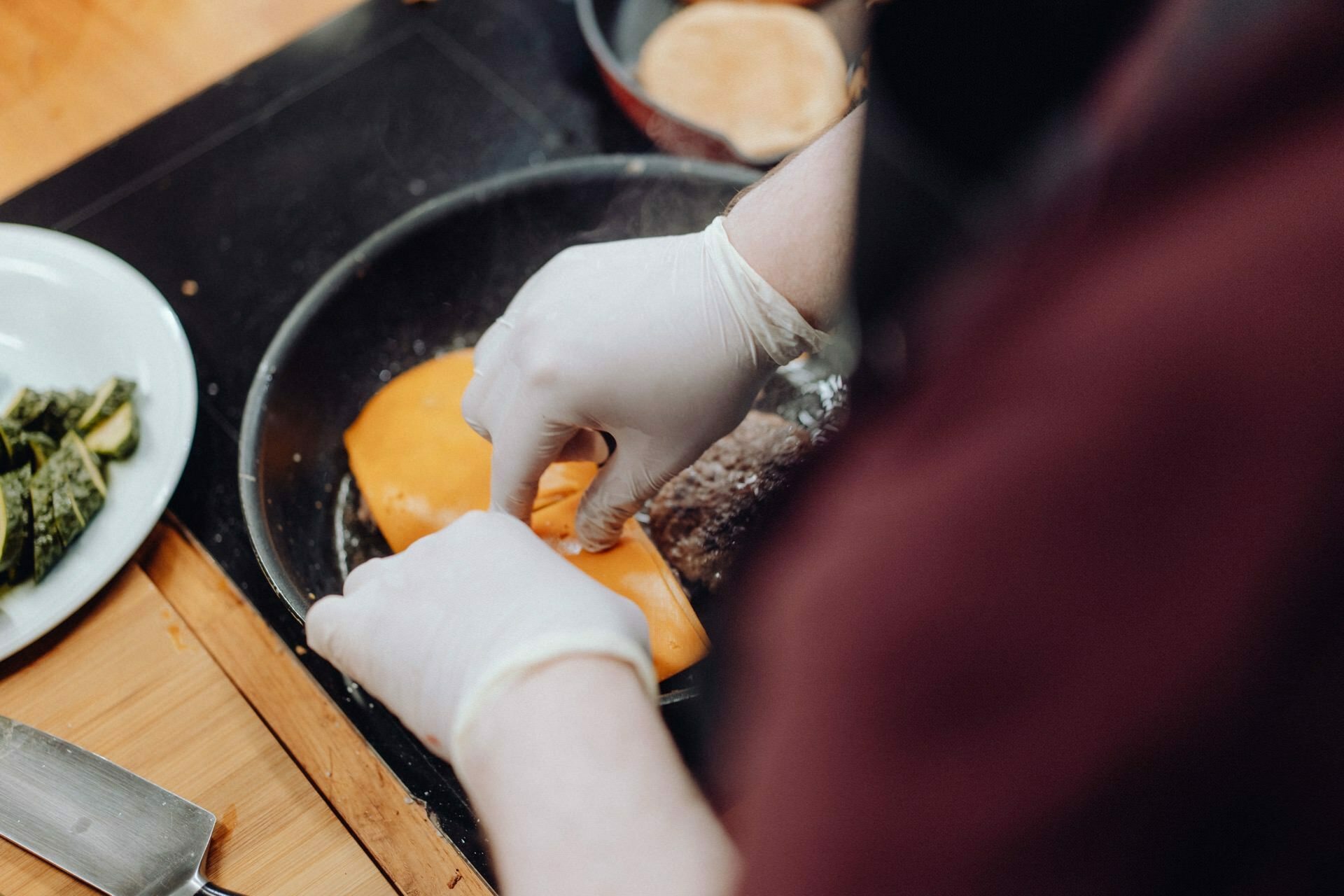 A person wearing white gloves puts cheese slices on a pate in a pan. Next to the vegetables on a white plate and a knife on a wooden cutting board, perfectly capturing the essence needed for event photography. 