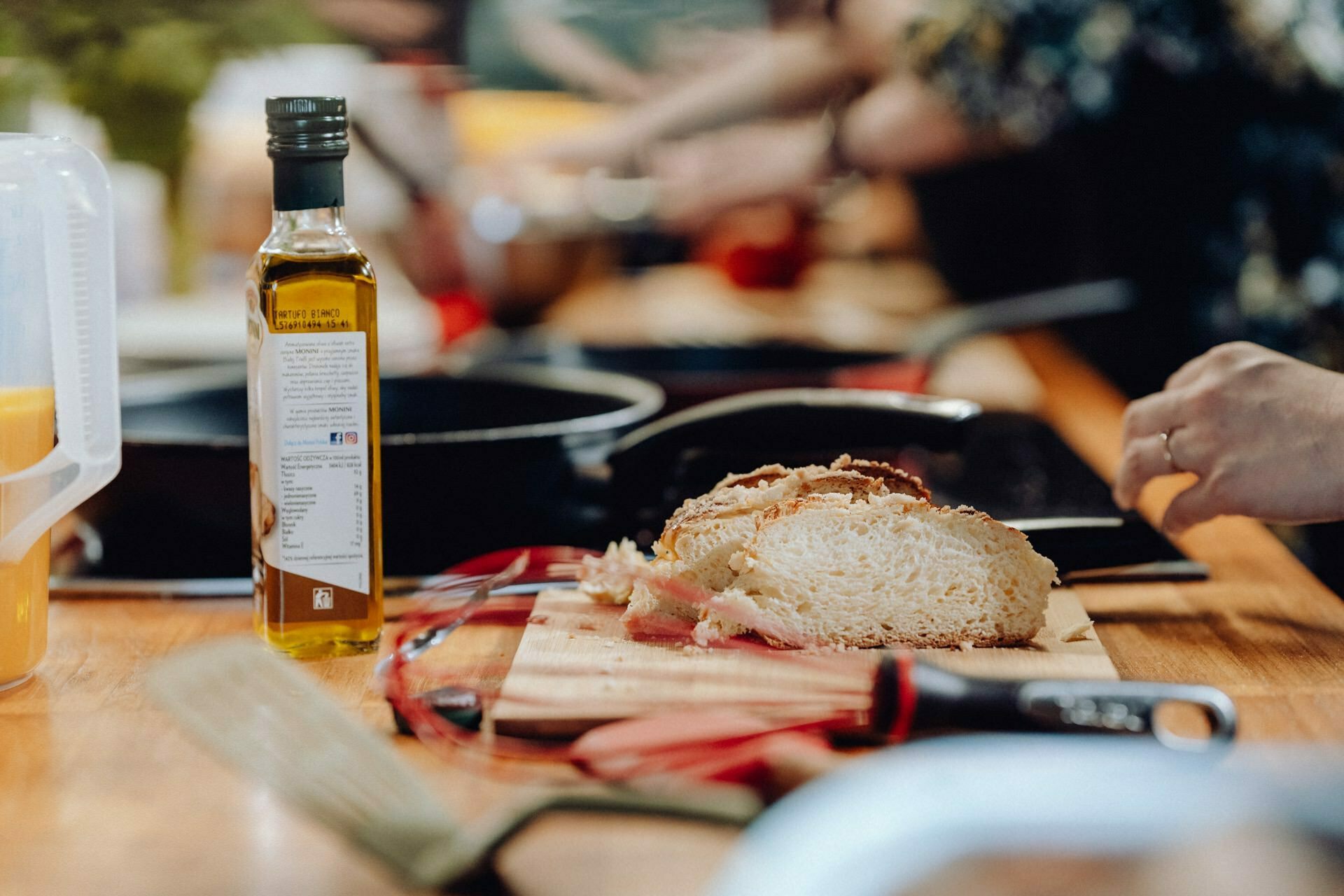 Close-up of a kitchen countertop with a cutting board on which lies sliced bread, a bottle of olive oil, a whisk, spatula and kitchen utensils. The blurry background depicts people engaged in cooking, capturing the vibrant essence of the photo event. 