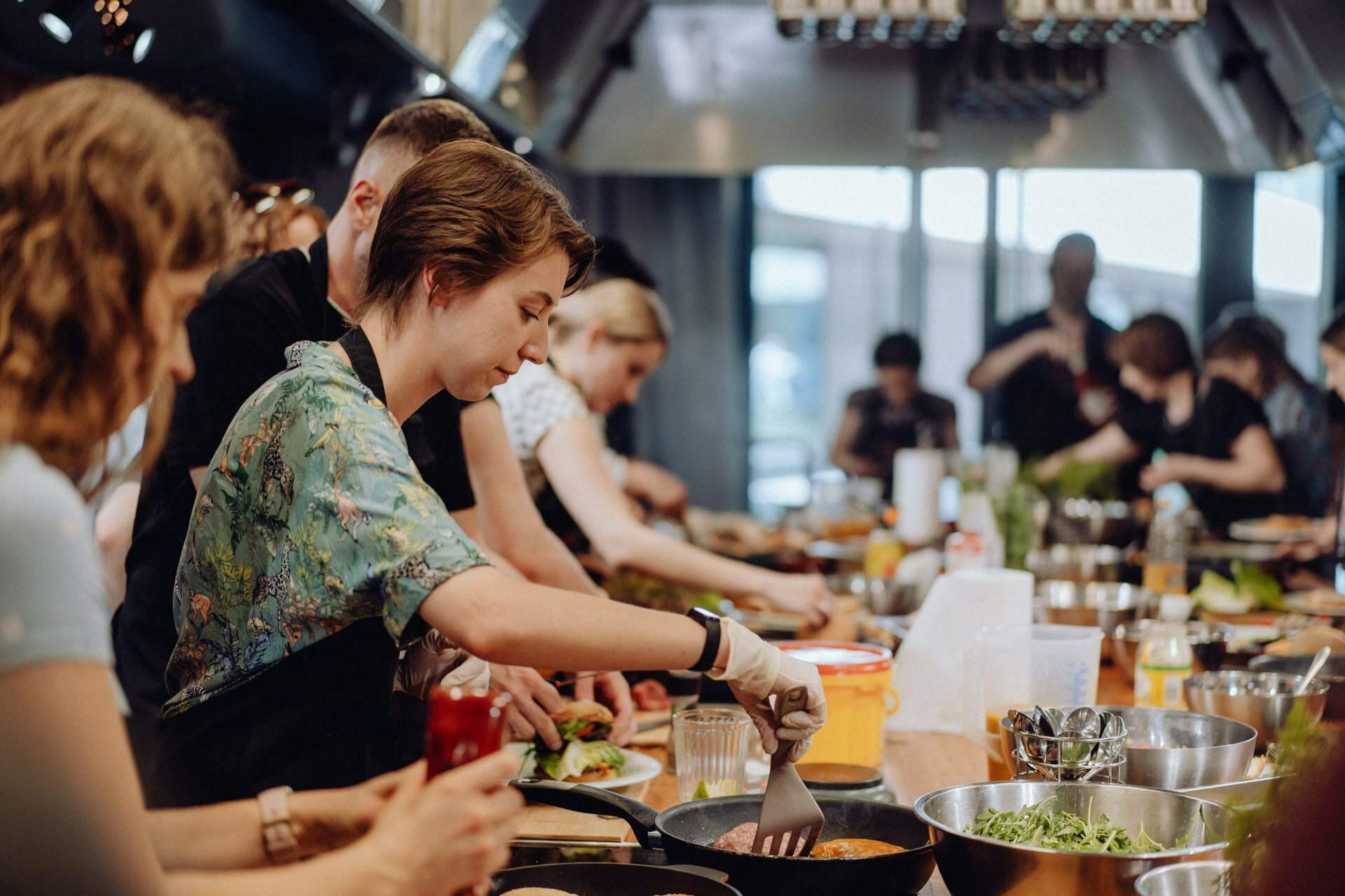 A group of people take part in a cooking class. In the foreground is a figure with short hair and a green floral shirt, stirring in a pan. Others in the background are preparing various dishes on a large wooden table filled with ingredients and cooking utensils, beautifully captured as part of an engaging photo essay of the events.  