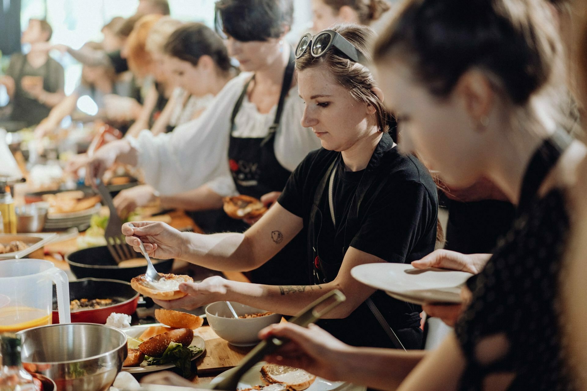 People are cooking and preparing food in a busy kitchen. Attention is focused on a woman in the foreground adding an ingredient to a dish, captured as part of an event photo essay by an event photographer Warsaw. Many other people are also busy cooking around her among the various utensils and ingredients spread out on the countertop.  