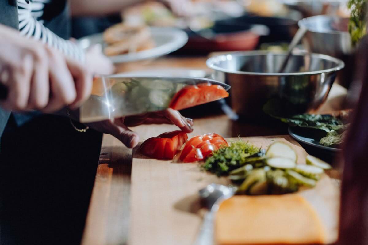 Zbliżenie osoby krojącej pomidory na drewnianej desce do krojenia w kuchni, idealne do fotorelacji z wydarzeń. Wokół deski do krojenia znajdują się różne składniki, w tym pokrojone w plasterki pikle, zioła i miski. W tle widoczna jest nieostra aktywność w kuchni, typowa dla fotografii eventowej.