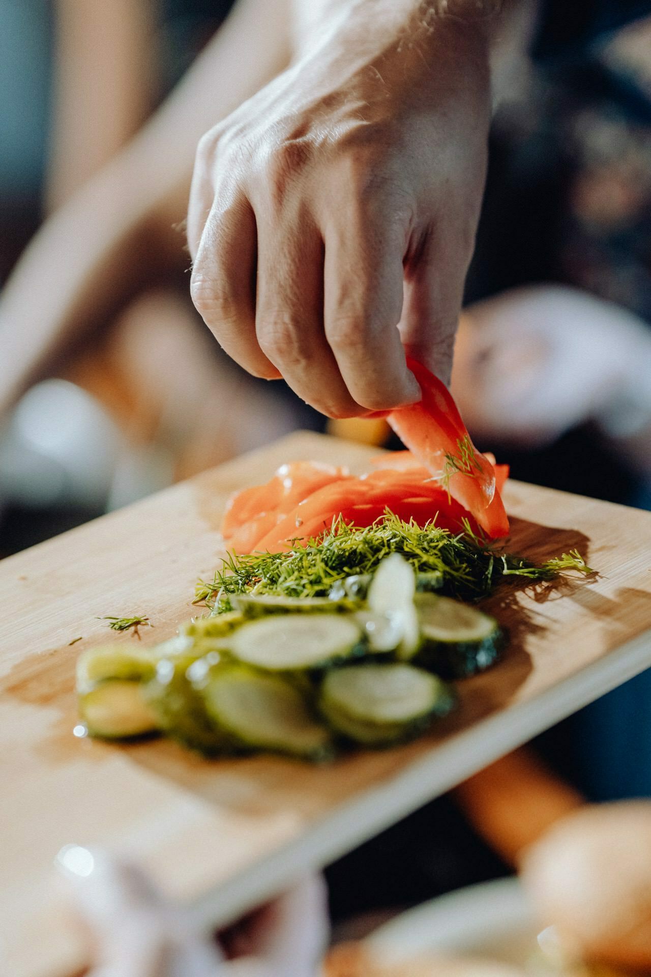 Close-up of a hand placing slices of red peppers on a wooden cutting board. Also on the board are sliced cucumbers and chopped dill, perfect details for event photography. The background is blurry and indistinct.  