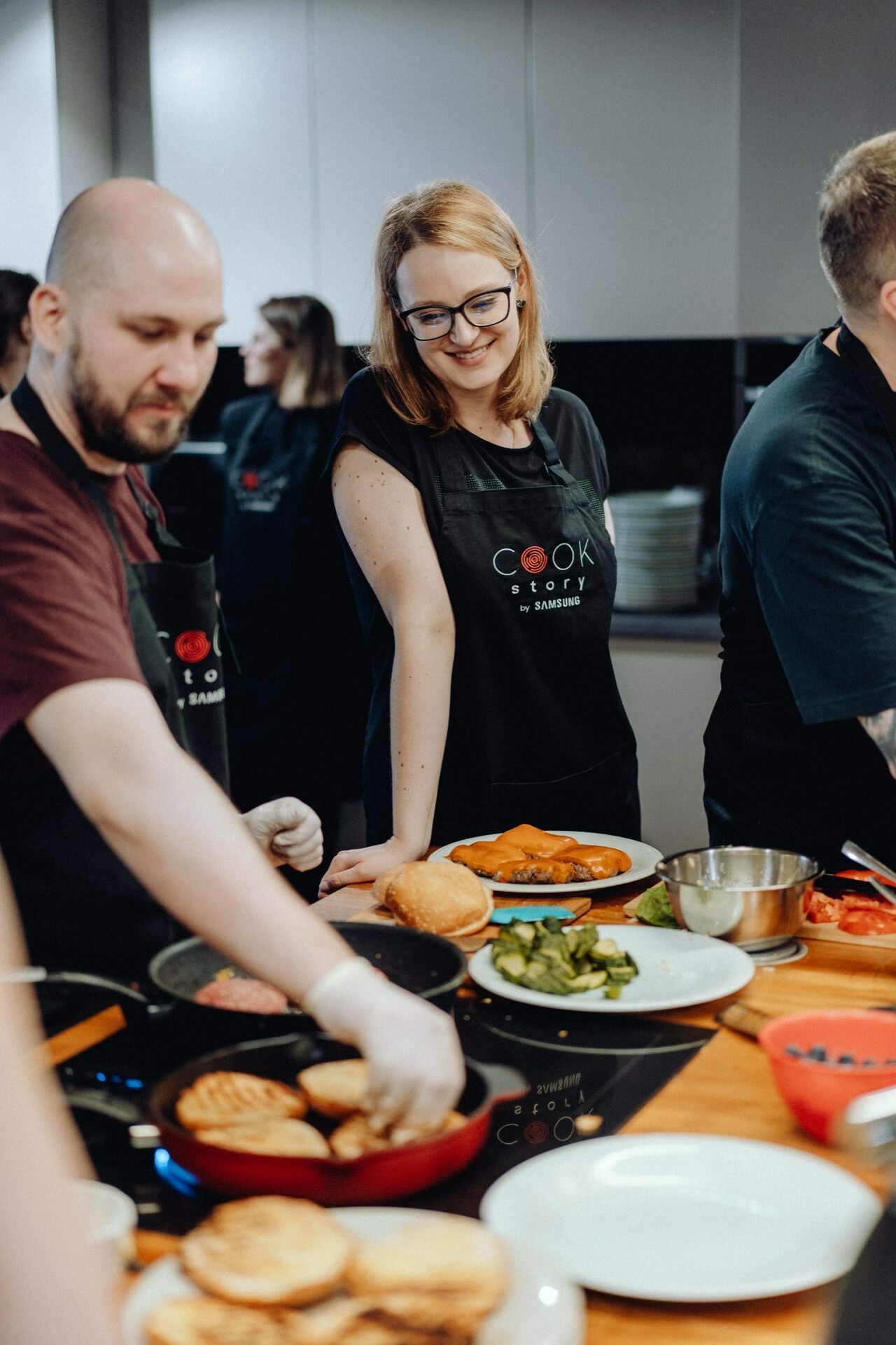 A group of people cooking together in the kitchen. A man grills food on the cooktop, while a smiling woman with glasses sitting next to him watches. Plates with prepared food and ingredients stand on the counter. Everyone in black aprons is capturing the delightful event photography.   