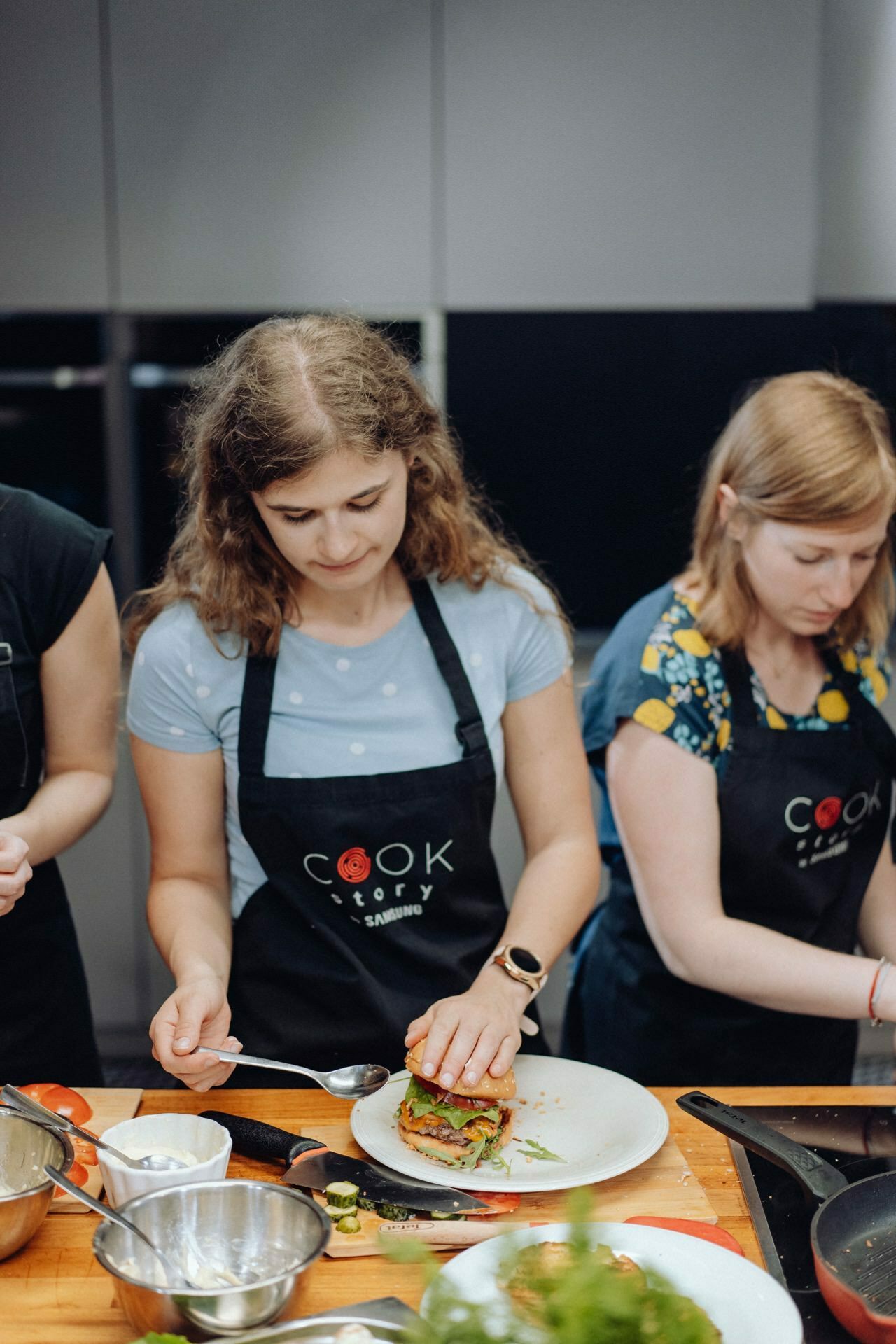 Two women wearing black aprons with the word "COOK" printed on them are preparing food in the kitchen. The woman on the left is using a spoon to arrange ingredients on a plate, while the woman on the right is focused on her task among various kitchen utensils. This scene could be part of an event photography showcasing culinary skills.  