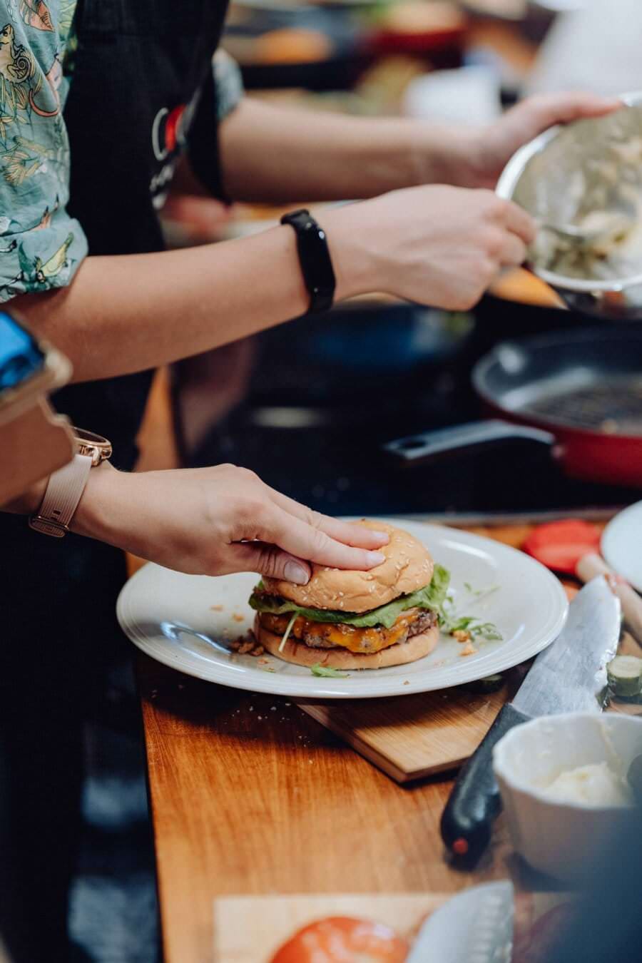 Osoba ubrana w czarny fartuch i smartwatch przygotowuje burgera na białym talerzu. Burger wypełniony jest sałatą, serem i pasztetem. Przybory kuchenne i składniki widoczne są na drewnianym blacie, a w tle pomaga im inna osoba – idealna scena do fotorelacji z wydarzeń.