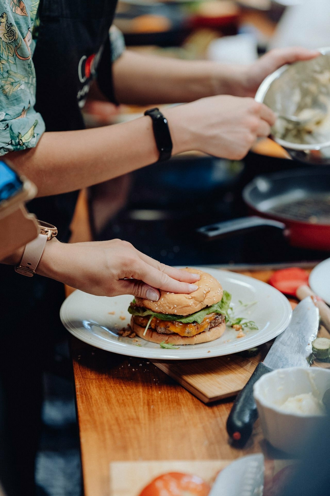 A person wearing a black apron and a smartwatch prepares a burger on a white plate. The burger is filled with lettuce, cheese and pate. The kitchen utensils and ingredients can be seen on a wooden countertop, while another person is helping them in the background - a perfect scene for a photo essay of the events.  