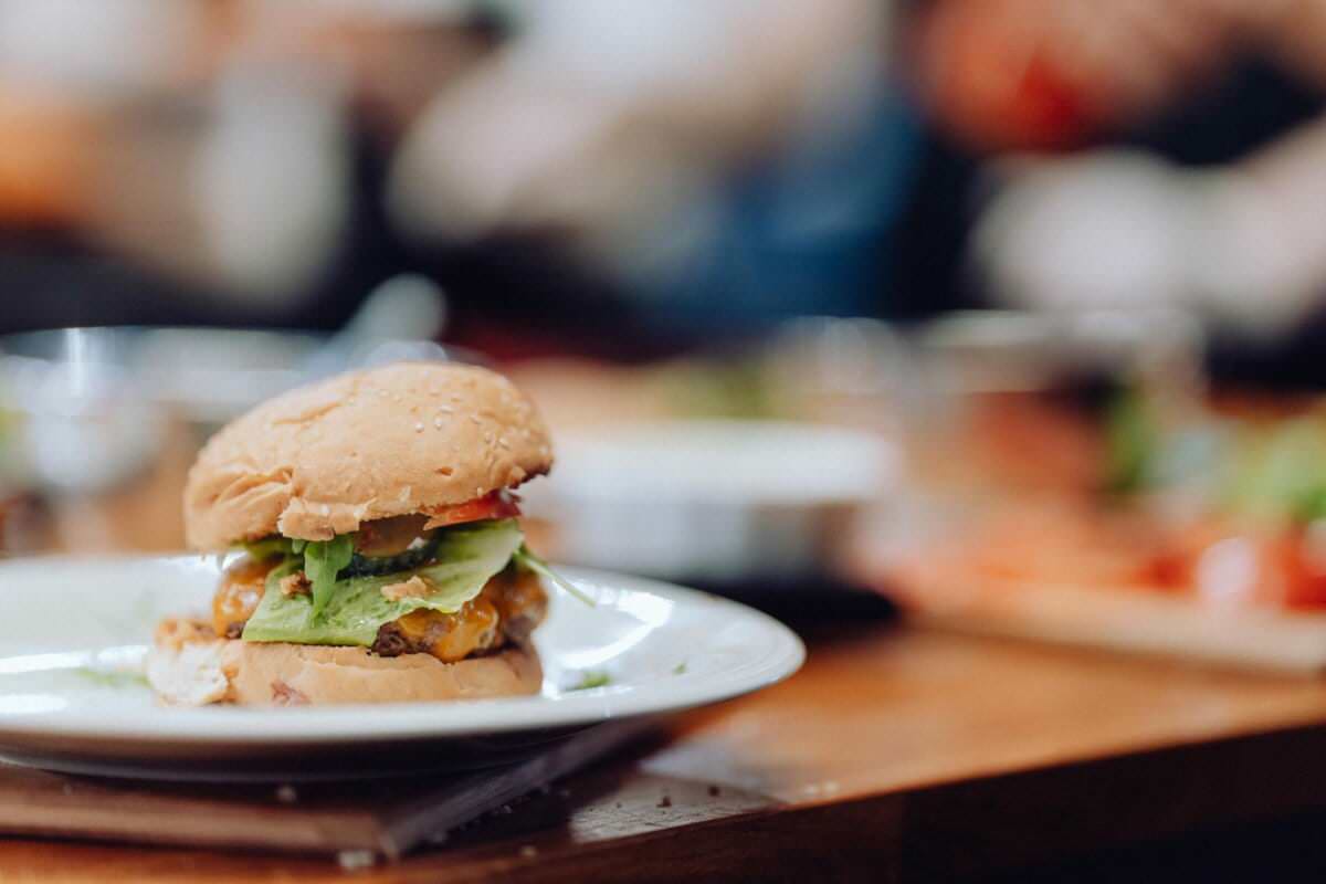 Zbliżenie cheeseburgera na białym talerzu, z bułką z sezamem, sałatą, pomidorem, serem i ewentualnie piklami i pasztecikiem burgerowym. Tło jest rozmyte, niczym fachowo uchwycona fotografia eventowa ustawiona na drewnianym stole z porozrzucanymi przedmiotami.