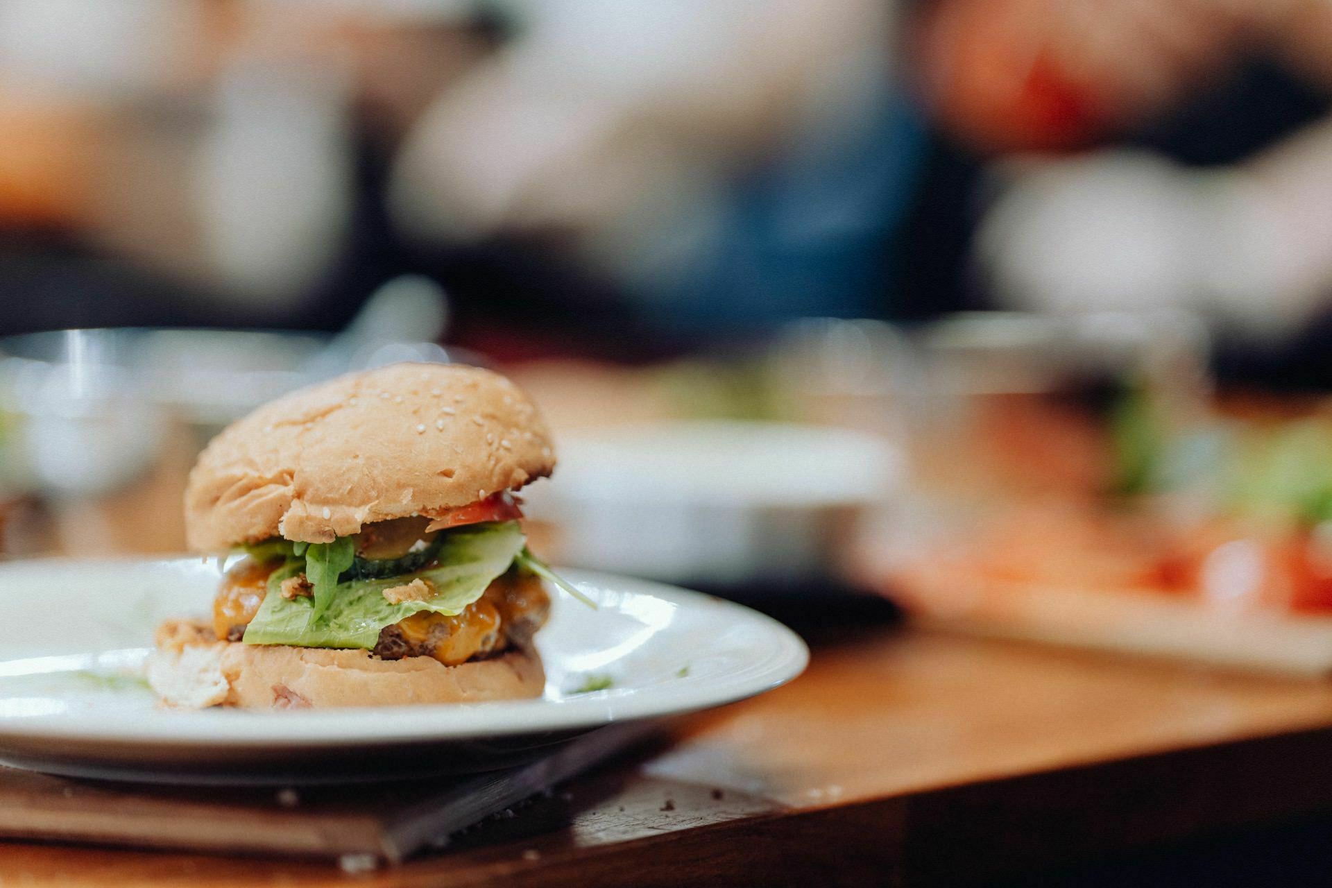 A close-up of a cheeseburger on a white plate, with a sesame seed bun, lettuce, tomato, cheese and possibly pickles and a burger patty. The background is blurred, like an expertly captured event photograph set on a wooden table with scattered objects. 