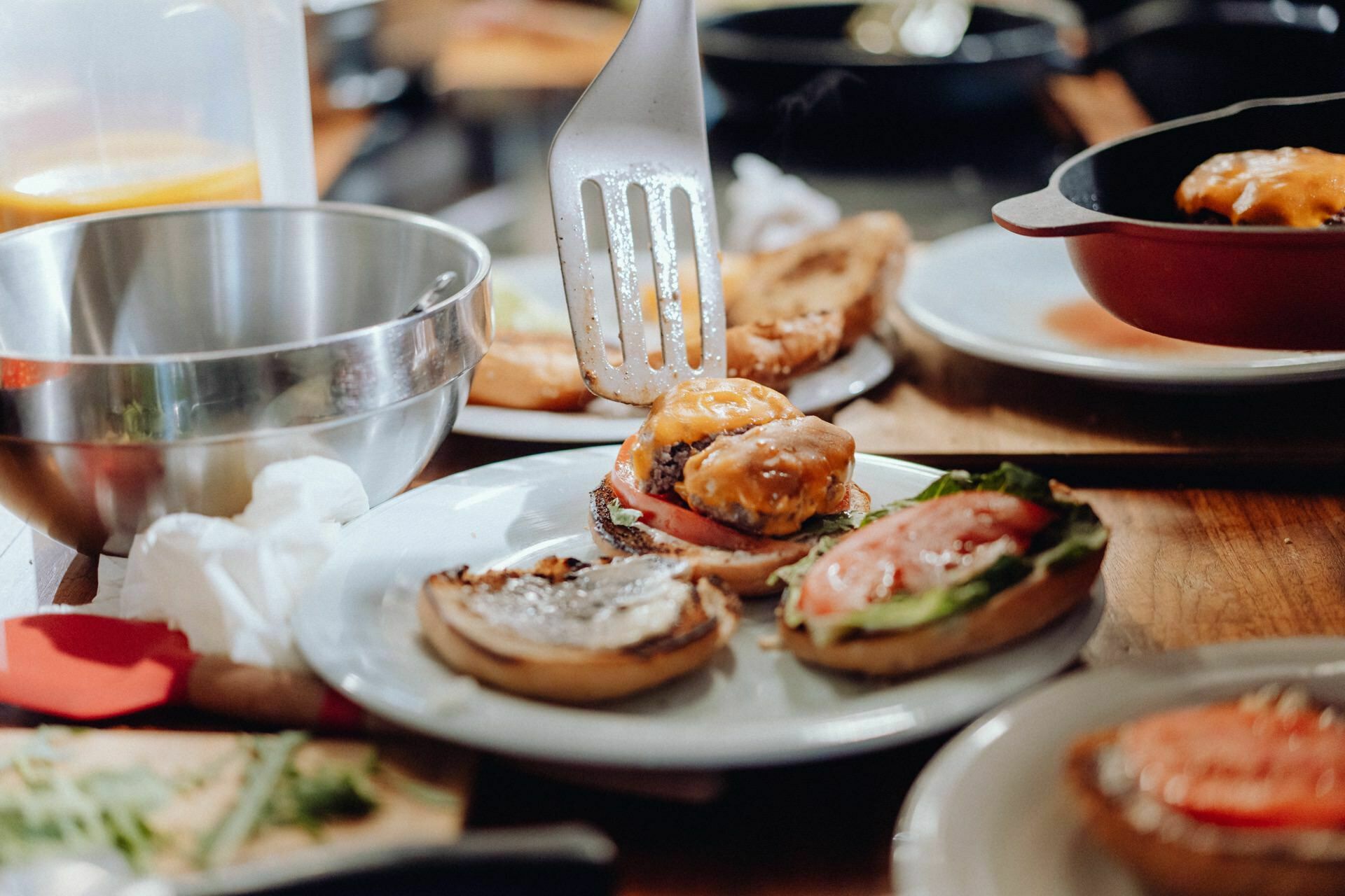 Close-up of a kitchen scene in which a spatula assembles a burger. A baked bun with melted cheese, beef patty, tomato and lettuce is placed on a plate. The wooden countertop shows various kitchen tools, ingredients and a metal mixing bowl; perfect for event photography enthusiasts.  