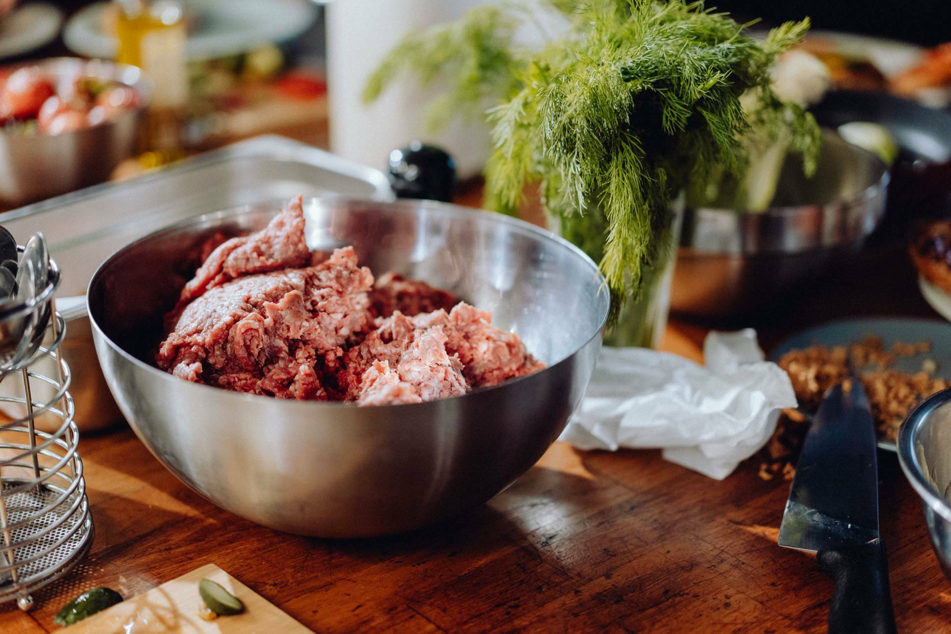 A large metal bowl filled with ground meat is placed on a wooden countertop, surrounded by various kitchen items, including a knife, paper towel, fresh dill and a metal food grinder. Other fuzzy ingredients and kitchen tools create the perfect setting for capturing event photography in the culinary heart of Warsaw. 