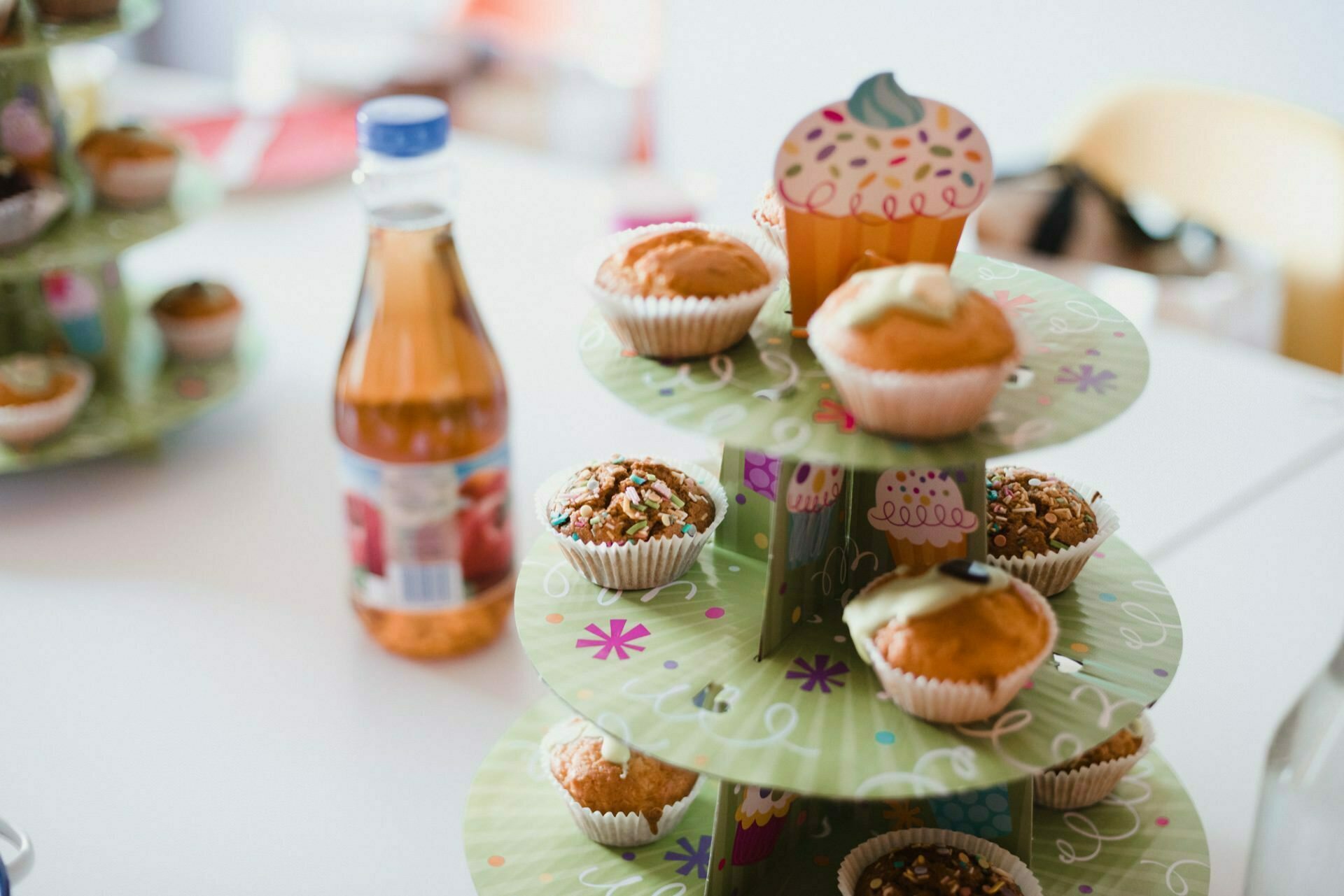 A multi-level stand with three tiers holds an assortment of decorated cupcakes in colorful packaging. A bottle of apple juice stands next to the stand. The background depicts a blurry interior, capturing the essence of a casual celebration in this stunning event photography.  