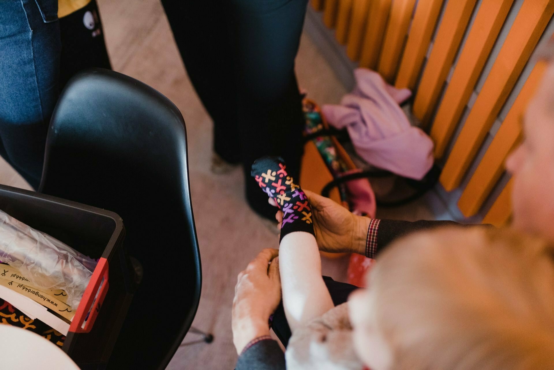 An adult helps a child put on colorful socks while sitting on a black chair. Various objects are visible in the background, including clothes on a wooden bench. Captured by an event photographer Warsaw, only the lower body and hands of the individuals are visible in this event photo.  