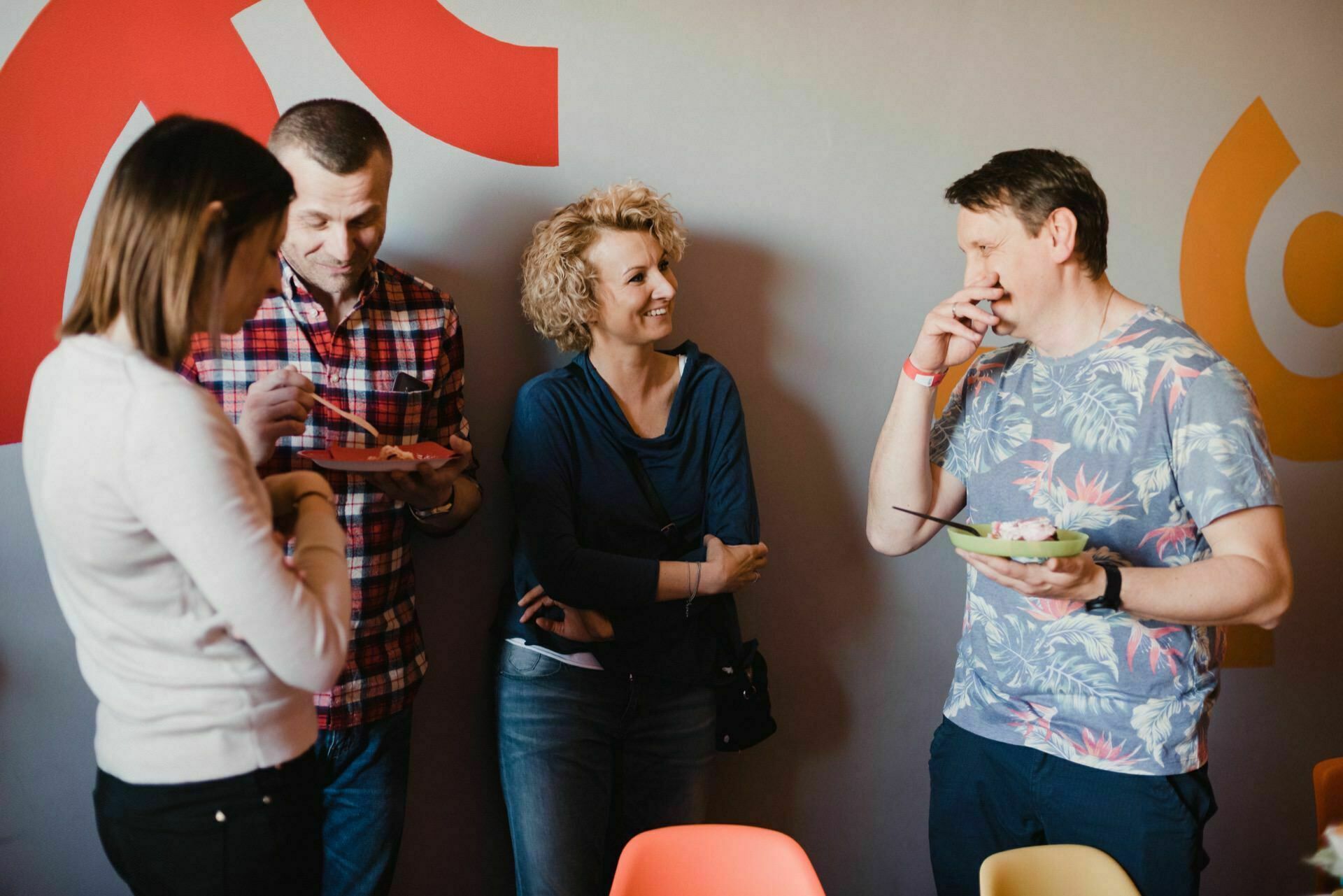 Four people gather at a party, two people are eating from plates. One person in a checkered shirt is talking to a woman in a blue sweater, another woman with a bob haircut is eating. A man in a floral shirt stands, laughing and eating against a colorful background. Photographs of events capture every moment.   