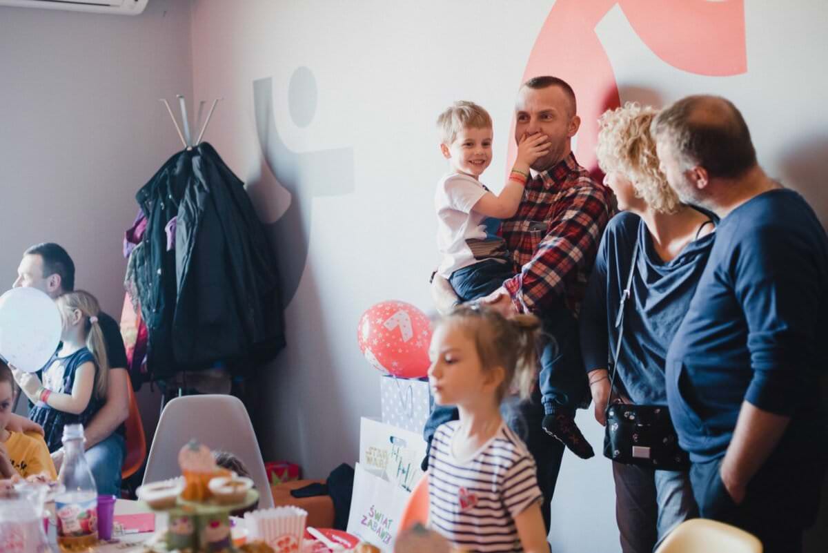Grupa dorosłych i dzieci zebrała się w pomieszczeniu, aby świętować. Mężczyzna trzyma uśmiechniętego chłopca, a obok bawi się dziewczynka w pasiastej koszuli. Dekoracje obejmują balony, przekąski i dekoracje świąteczne na stołach i ścianach. W tle na wieszaku wiszą płaszcze – pięknie uchwycone w ramach fotorelacji z wydarzeń.