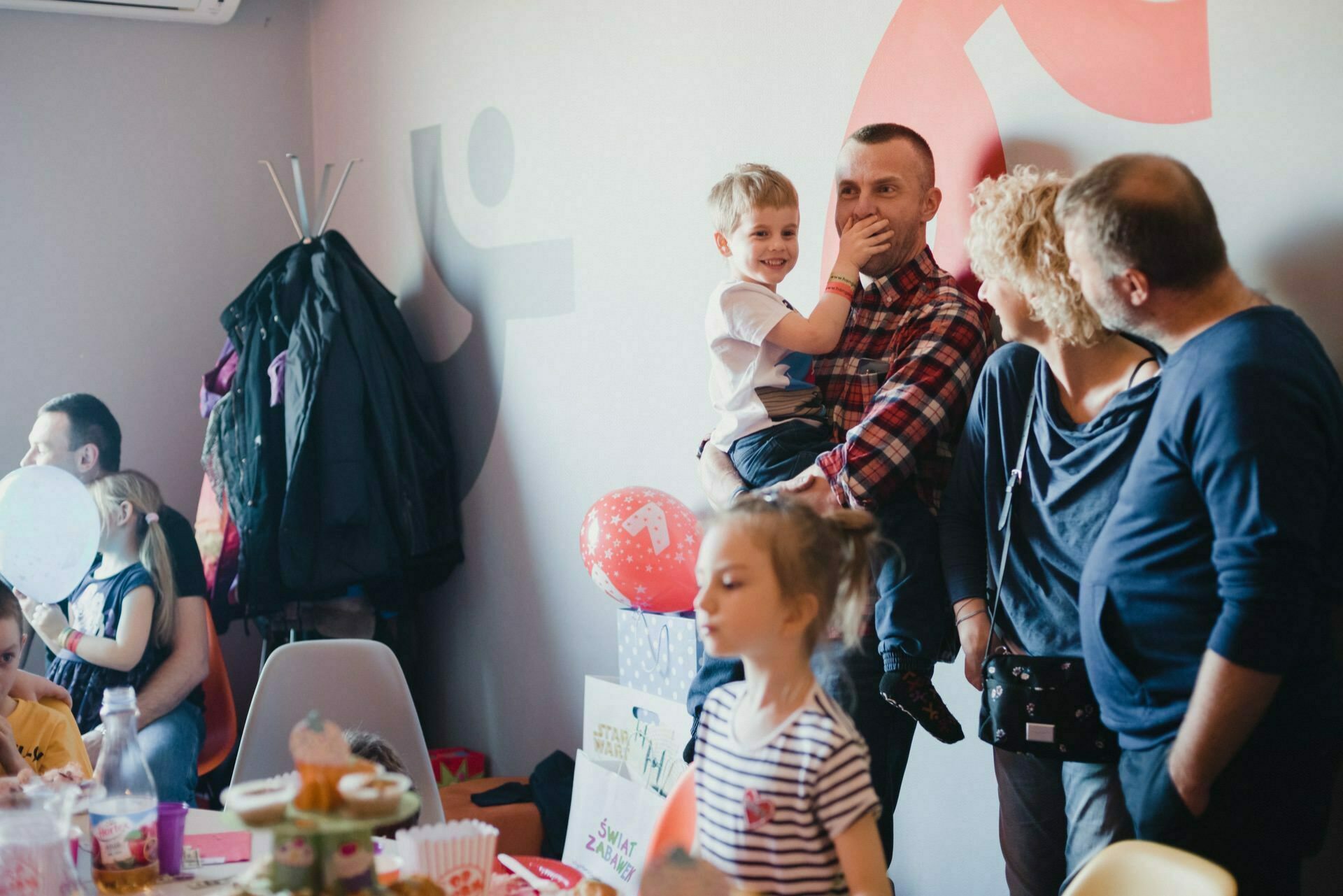 A group of adults and children have gathered in a room to celebrate. A man holds a smiling boy, while a girl in a striped shirt plays next to him. Decorations include balloons, snacks and Christmas decorations on the tables and walls. In the background, coats hang on a coat rack - beautifully captured as part of the event photo coverage.   