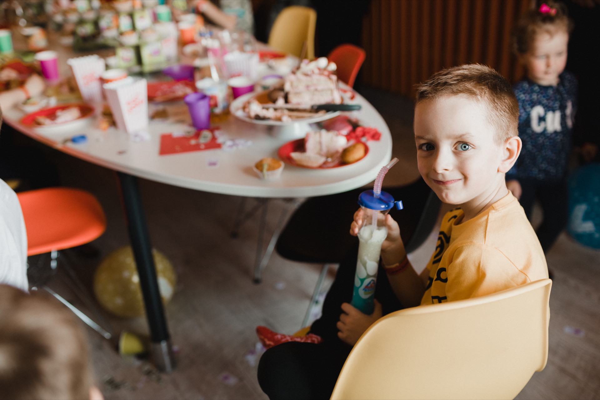 A young boy with short brown hair smiles and holds a drink with a straw while sitting on a yellow chair at a table full of snacks and party gadgets. Other children can be seen in the background, capturing the lively essence of the gathering, perfect for event photography. 