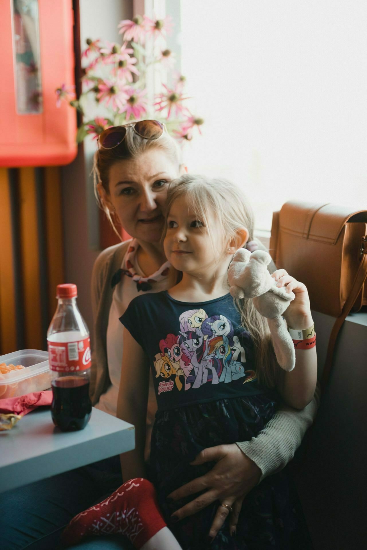 A woman with sunglasses on her head sits next to a young girl holding a plush toy. The girl is dressed in a dress with colorful animated characters. On the table stands a soda bottle with a red label and a plastic container of food. In the background, pink flowers capture a joyful moment, perfect for a photo event.   