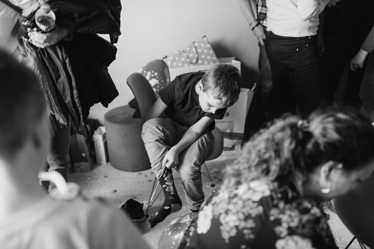 Czarno-białe zdjęcie młodego chłopca siedzącego na krześle na zgromadzeniu lub imprezie. Patrzy w dół i wydaje się być głęboko zamyślony. Wokół są ludzie, ale ich twarzy nie widać. W tle widać balony i dekoracje, typowe dla fotografii eventowej oddającej istotę chwili.