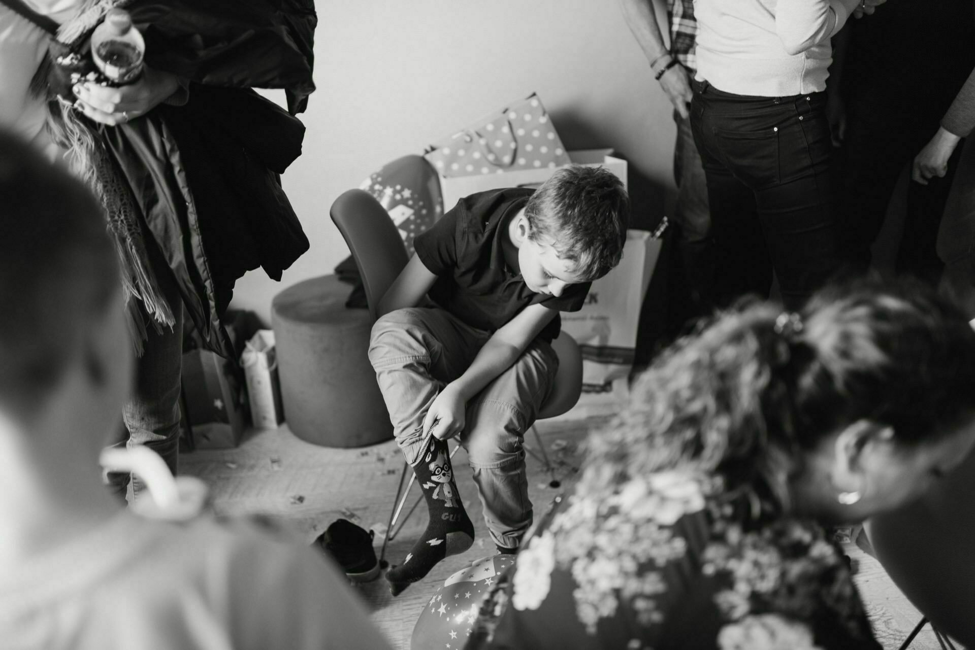 Black and white photo of a young boy sitting on a chair at a gathering or event. He is looking down and appears to be deep in thought. There are people around, but their faces are not visible. Balloons and decorations can be seen in the background, typical of event photography capturing the essence of the moment.   