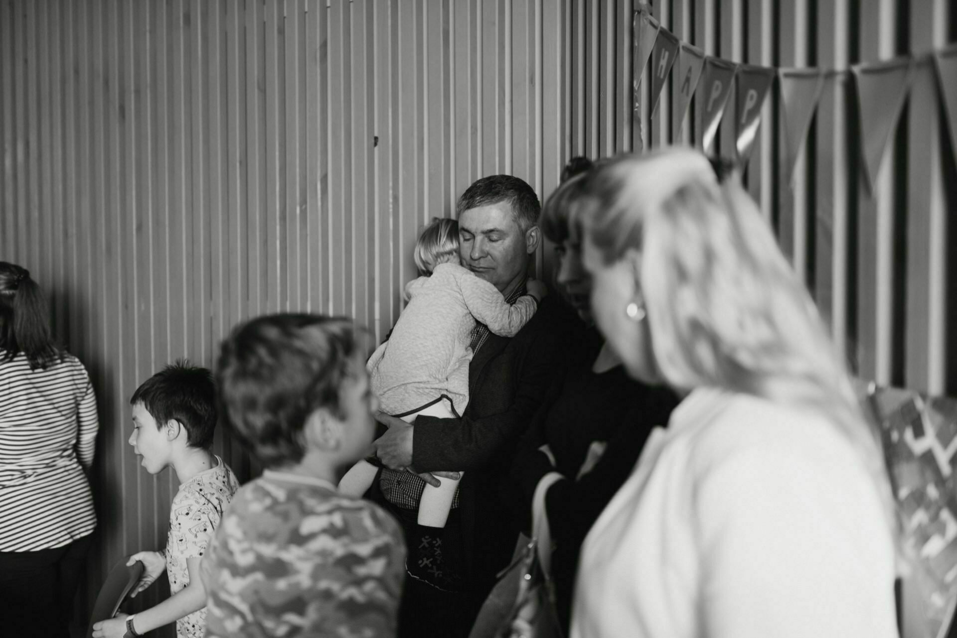 Black and white photo of a group of people, including several children. In the center, a man is hugging a child. There are other children nearby, and the woman in the foreground is facing to the side. A flag hangs on the wall in the background, capturing a touching moment from this event photograph.   